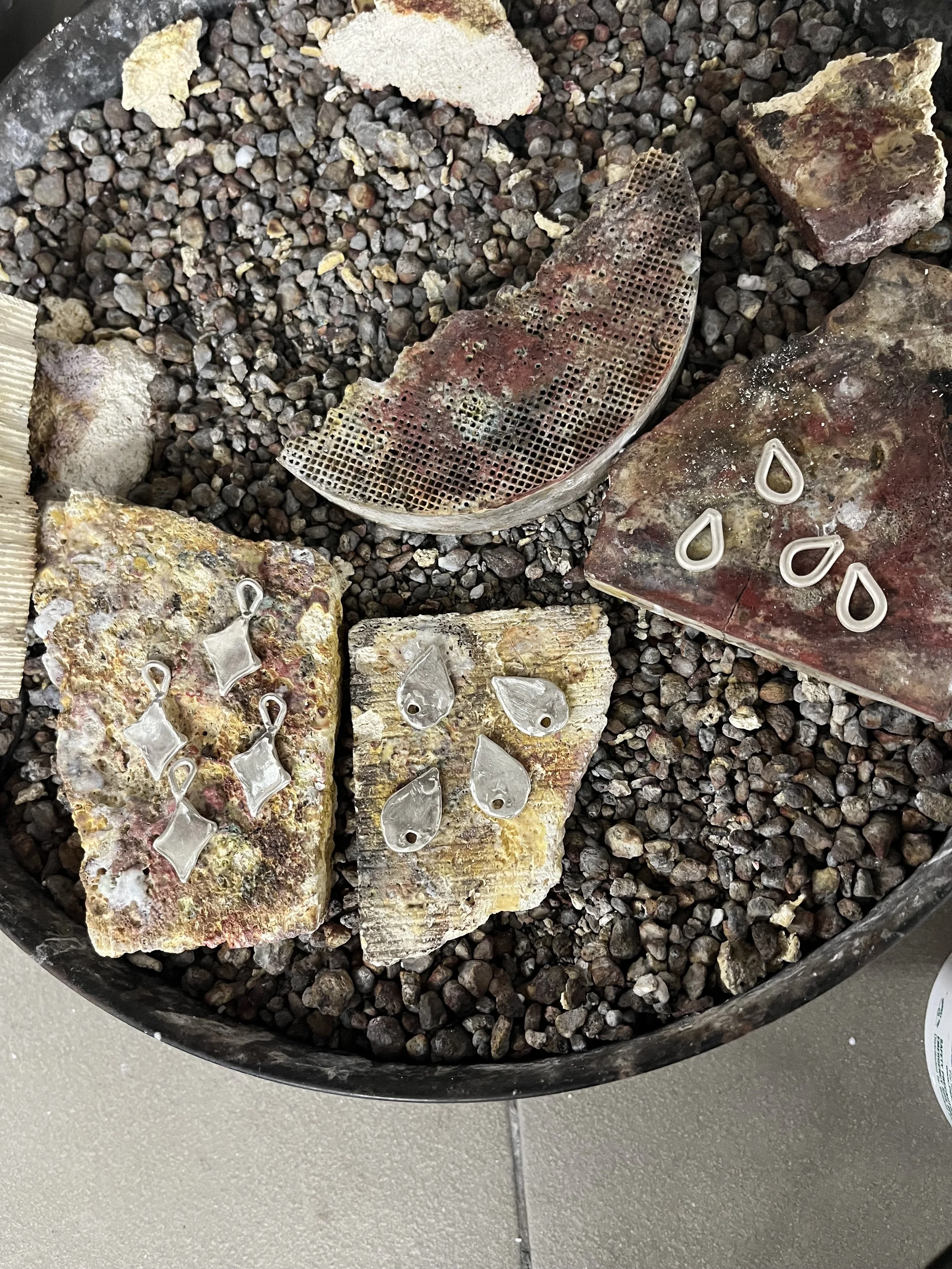 Various rocks with embedded glass pieces and small metal hooks placed on small gravel stones, possibly part of a jewelry or craft project.