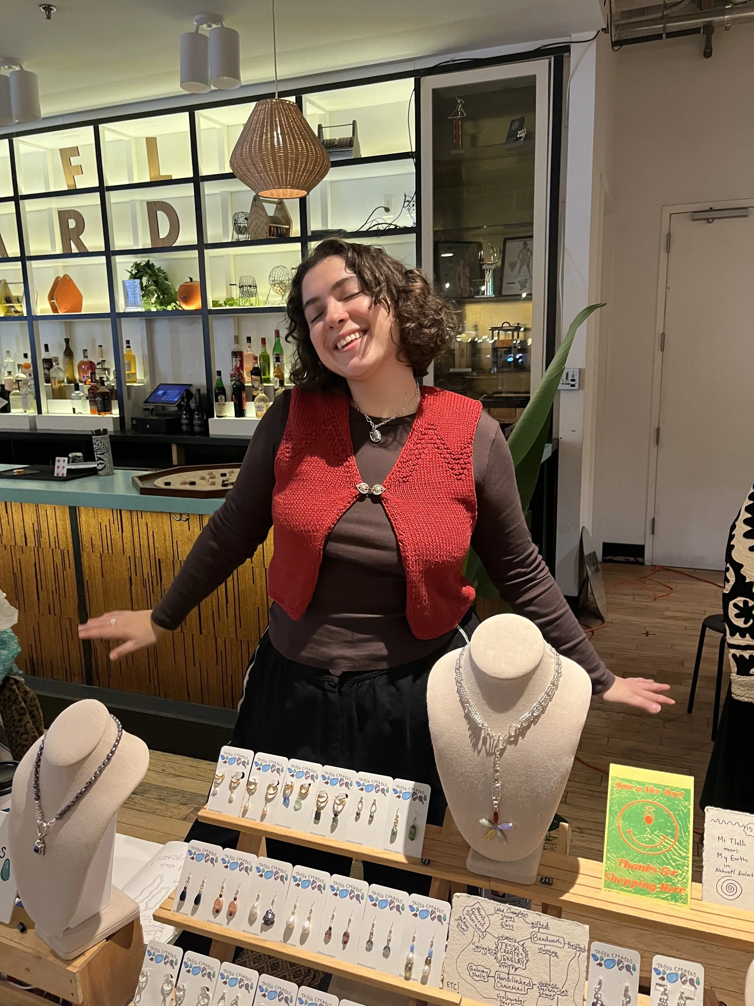 A woman with curly hair and a red vest smiling at a jewelry display table in a store or market. The table features necklaces and earrings on display busts and cards, with a background of shelves filled with decorative items and bottles.