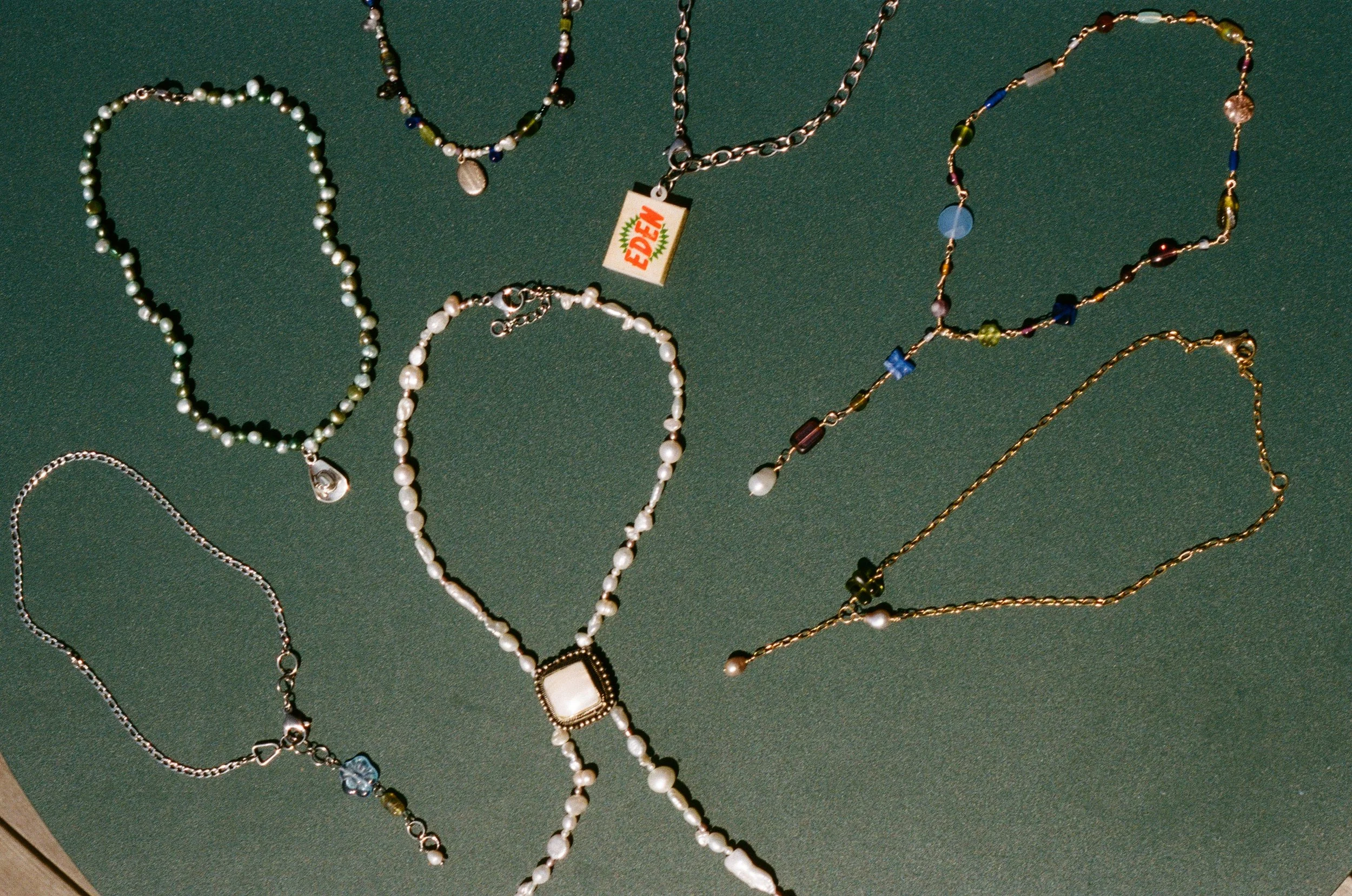 A collection of necklaces and bracelets displayed on a green surface. The jewelry includes beaded and chain styles, with some featuring pendants or charms.