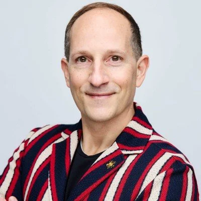 A man with short, dark hair and fair skin wearing a black shirt and a striped blazer with red, white, and blue colors, smiling at the camera against a light gray background.