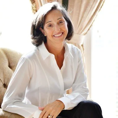 A smiling woman with shoulder-length dark hair, wearing a white blouse, sitting on a beige sofa in a sunlit room with beige curtains.