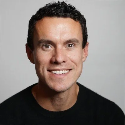A smiling man with short dark curly hair wearing a black shirt against a light gray background.