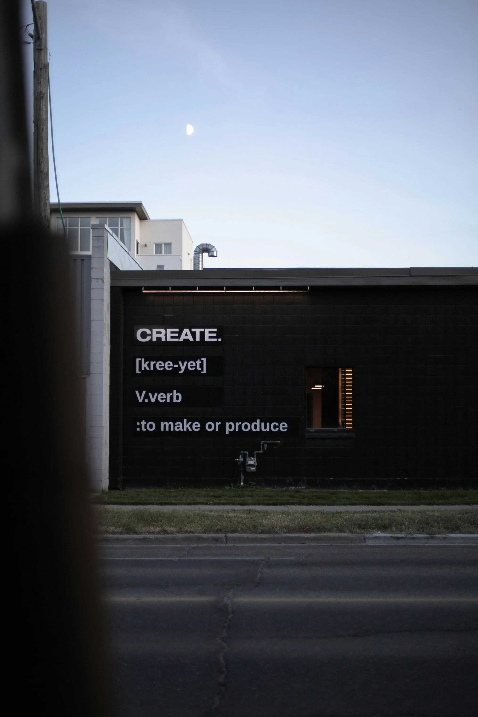 A black building with a sign defining the word 'CREATE,' to make or produce. The sign includes the word's pronunciation and part of speech, and the background shows the sky with a moon.
