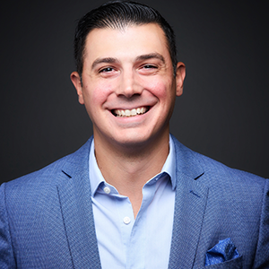 A smiling man with short dark hair, wearing a blue suit jacket and a light blue shirt against a dark background.