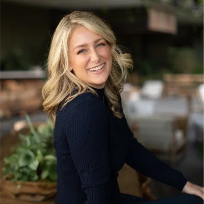 A young woman with blonde hair smiling, wearing a dark sweater, outdoors in a garden or park setting