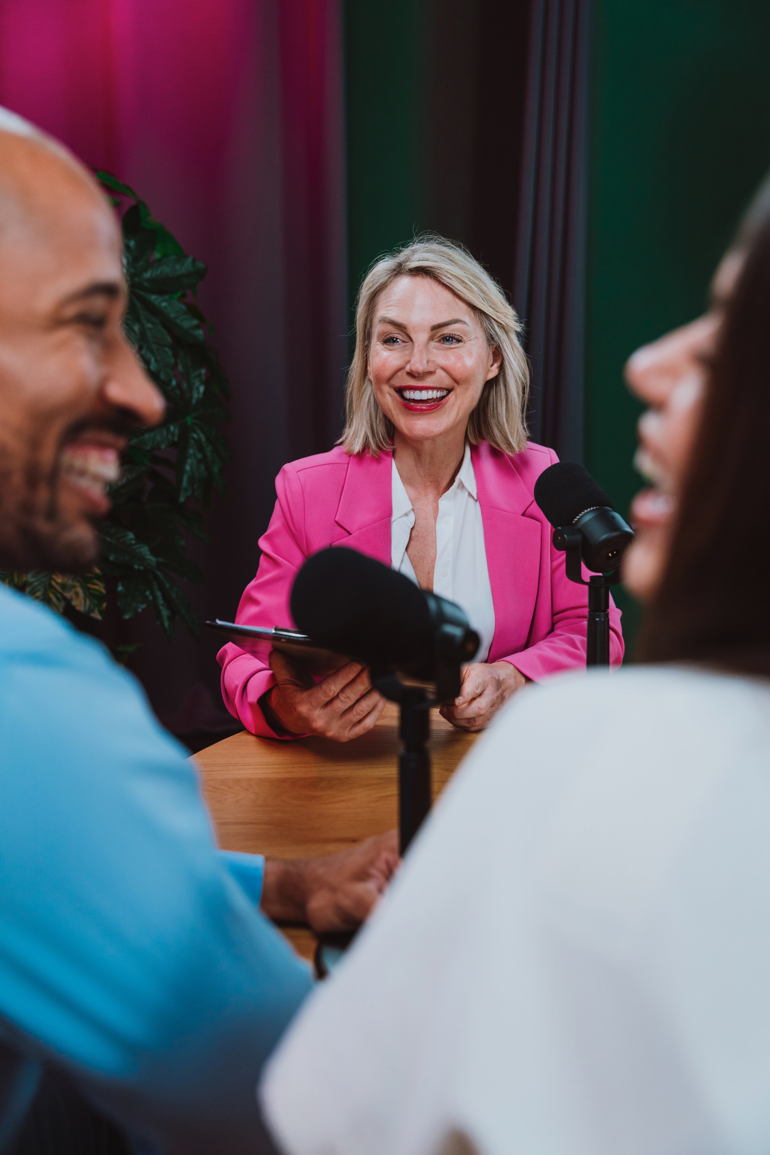 A woman in a pink blazer conducting a podcast with two guests, smiling, with microphones set up in front of them.
