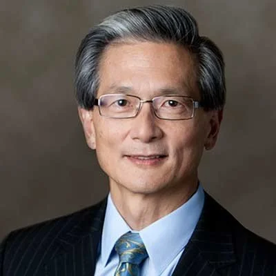 A professional headshot of an older Asian man with glasses, gray hair, wearing a dark suit, light blue shirt, and patterned tie, against a neutral background.