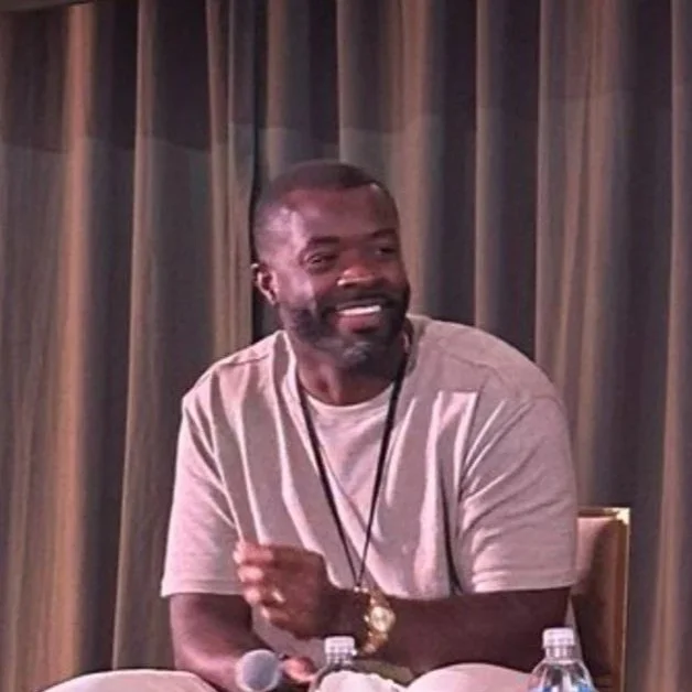 A man with a beard sitting at a table with a microphone, water bottles, and a laptop, smiling, in front of a curtain.