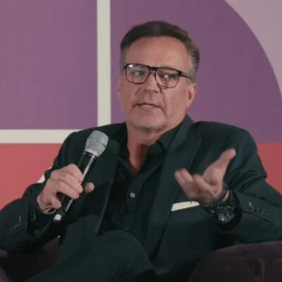 A man with short gray hair, glasses, and a black blazer holding a microphone and gesturing with his left hand during a discussion or interview.