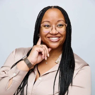 A smiling woman with glasses and braided hair, wearing a beige shirt, posing with her hand on her chin against a light background.