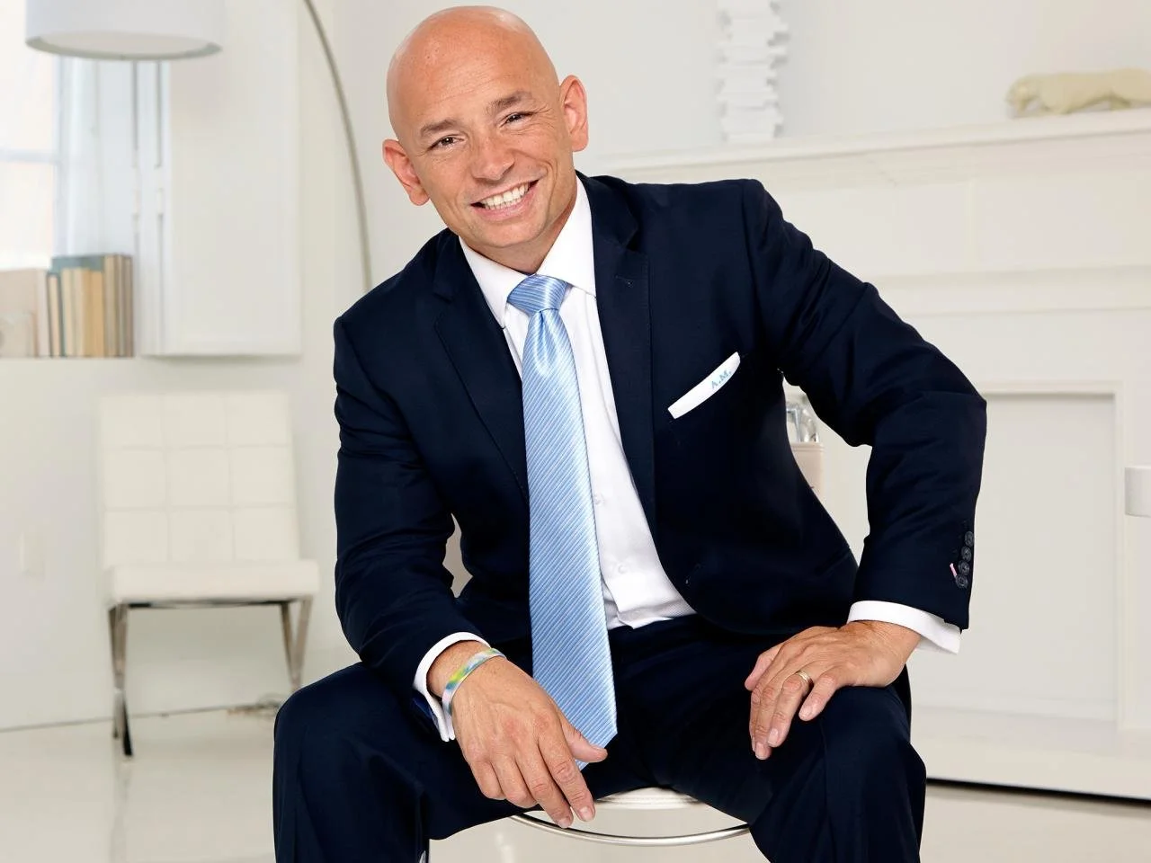 A smiling bald man in a dark suit and light blue tie, seated in a bright, modern room with white walls and minimal decor.