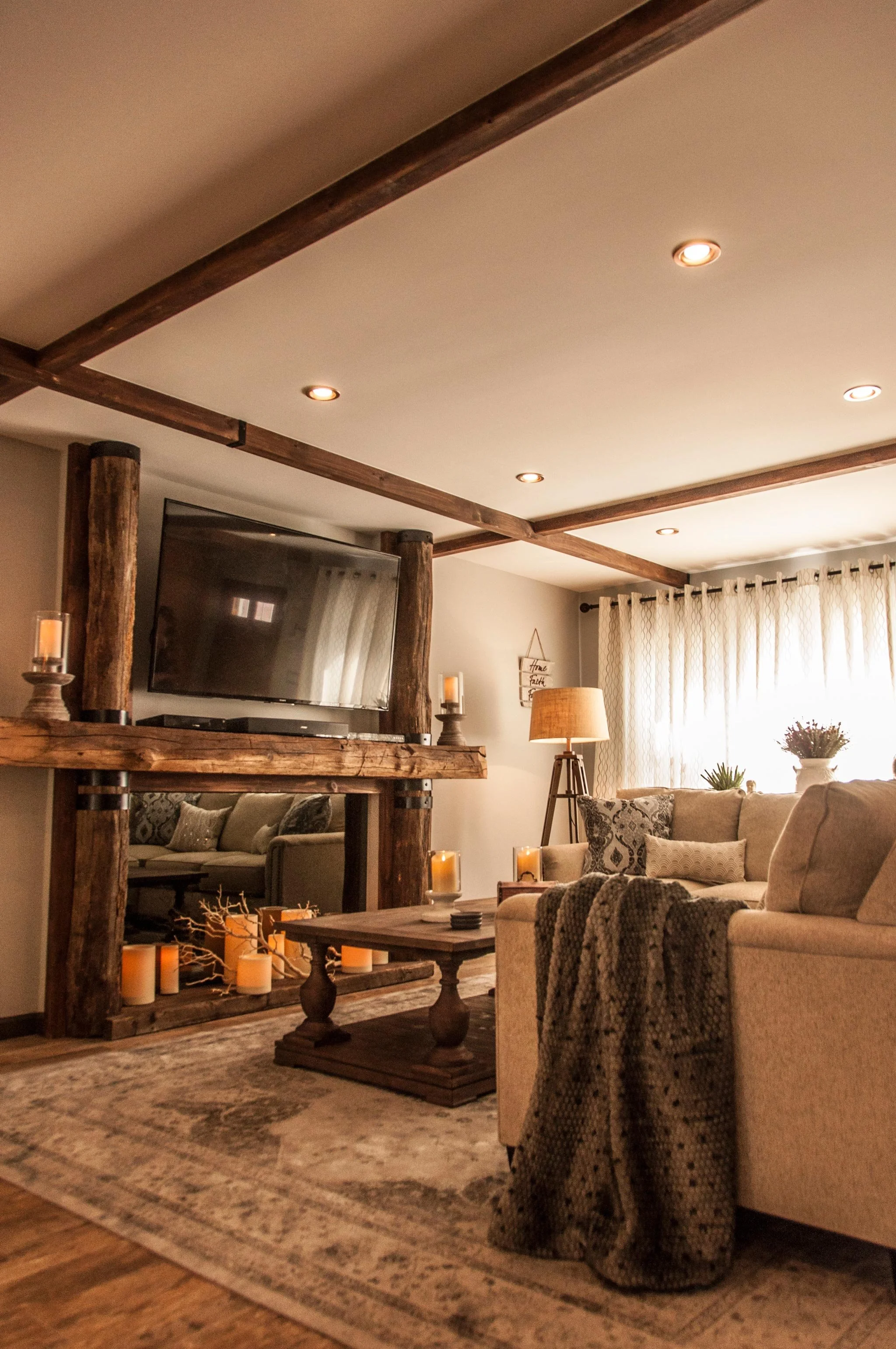 Cozy living room with beige sofas, wooden coffee table, and a rustic wooden fireplace mantel decorated with candles. There is a large window with curtains, a floor lamp, and decorative plants.