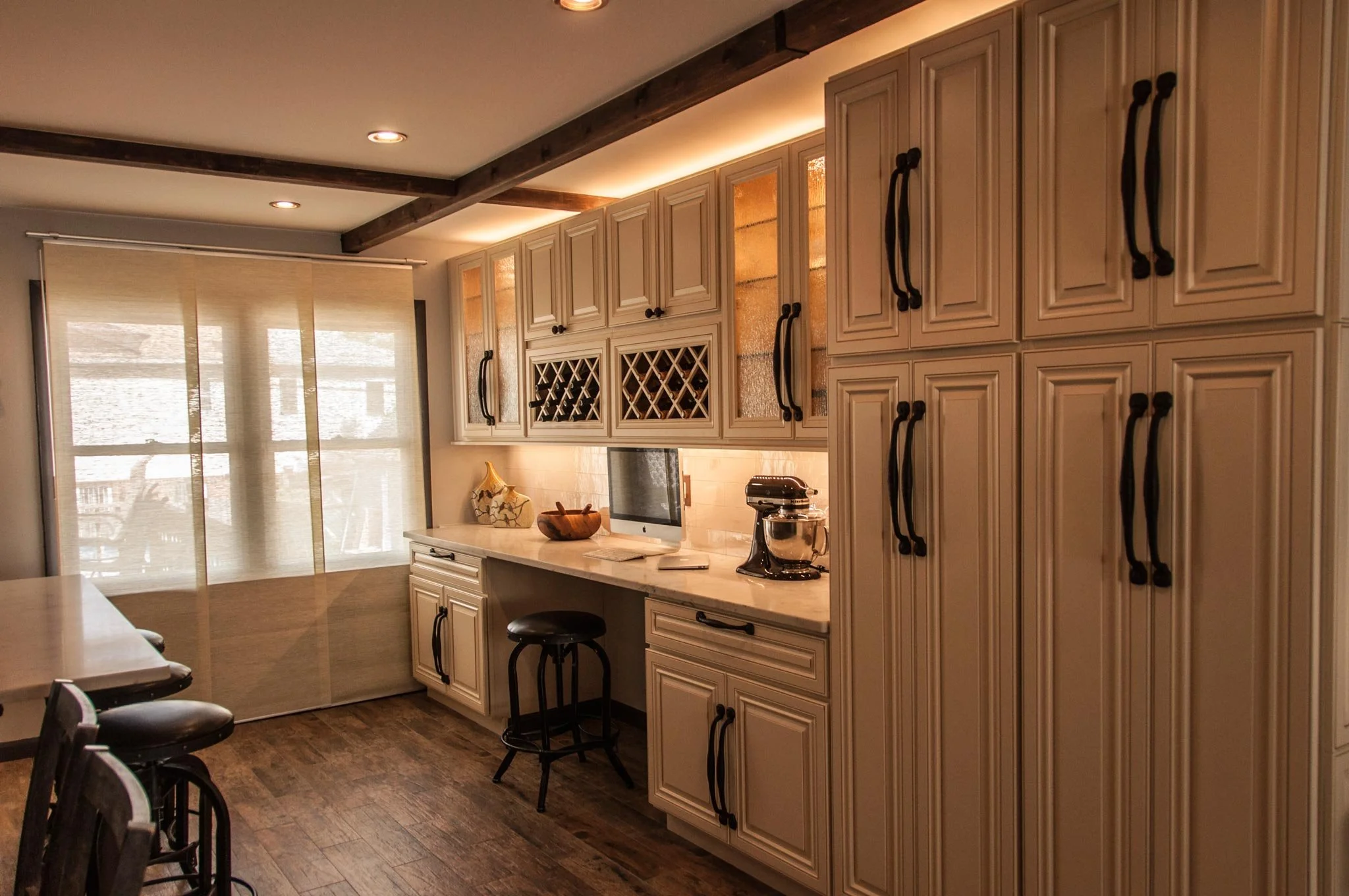 A kitchen with beige cabinets, black handles, a small TV, a stand mixer, and wine storage, with a wooden floor and a window with blinds.