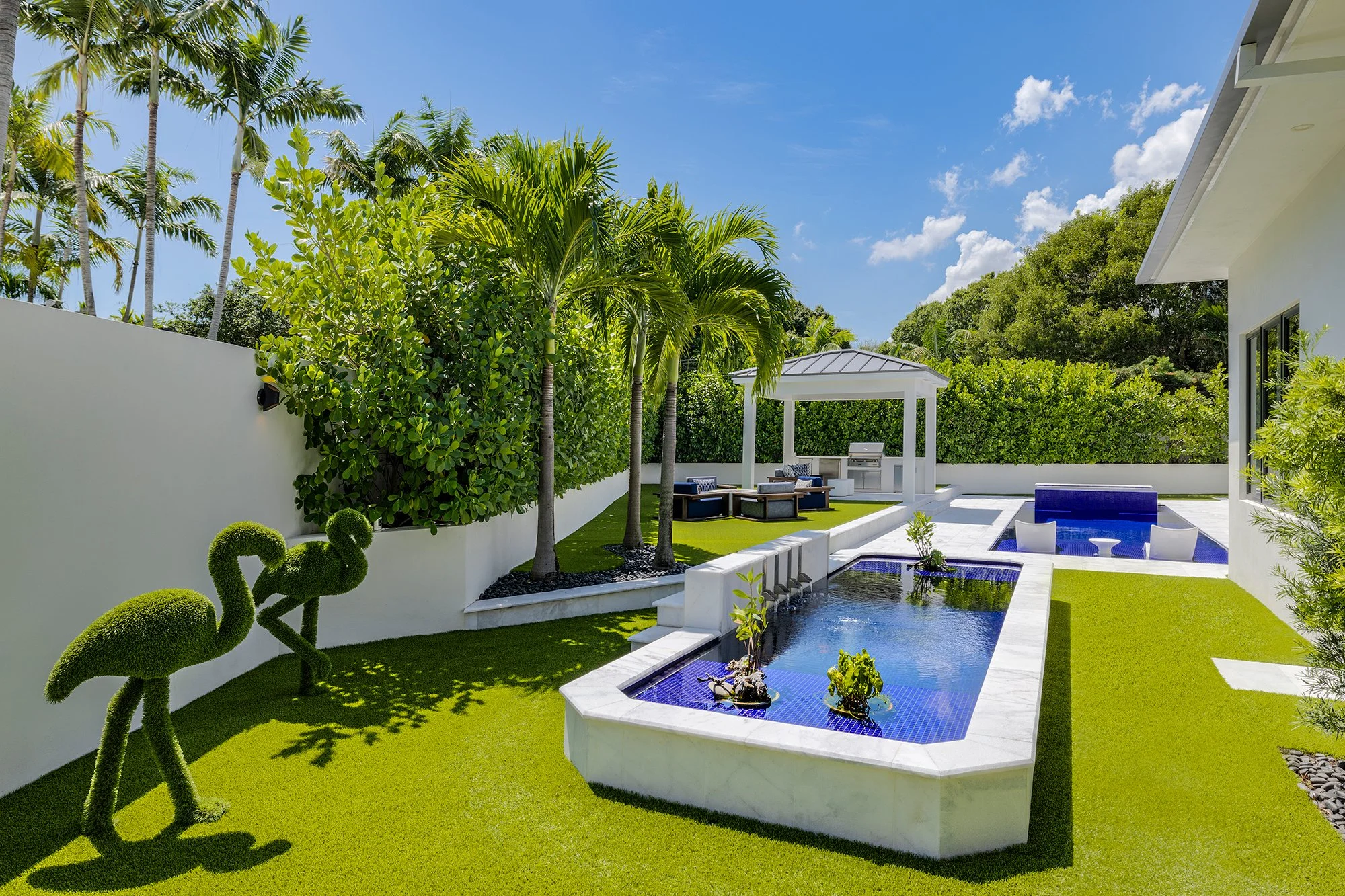 A modern backyard with a small pool, lush green grass, palm trees, a gazebo, outdoor seating, a barbecue grill, and decorative flamingo sculptures, under a bright blue sky.