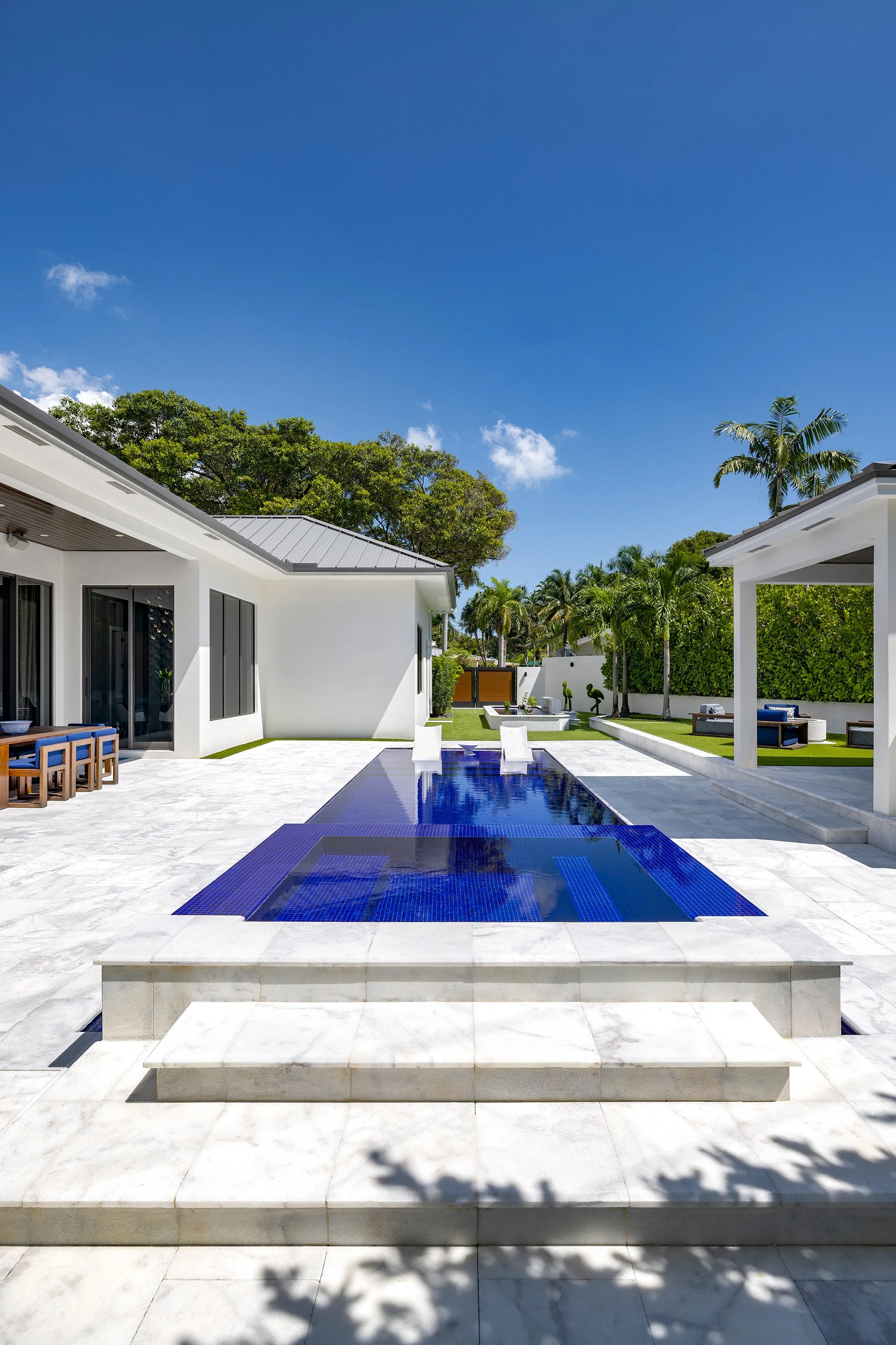 Modern backyard with a rectangular pool, white marble tiles, and outdoor seating area under a white pergola. Green trees and palm trees surround the yard, with a bright blue sky overhead.