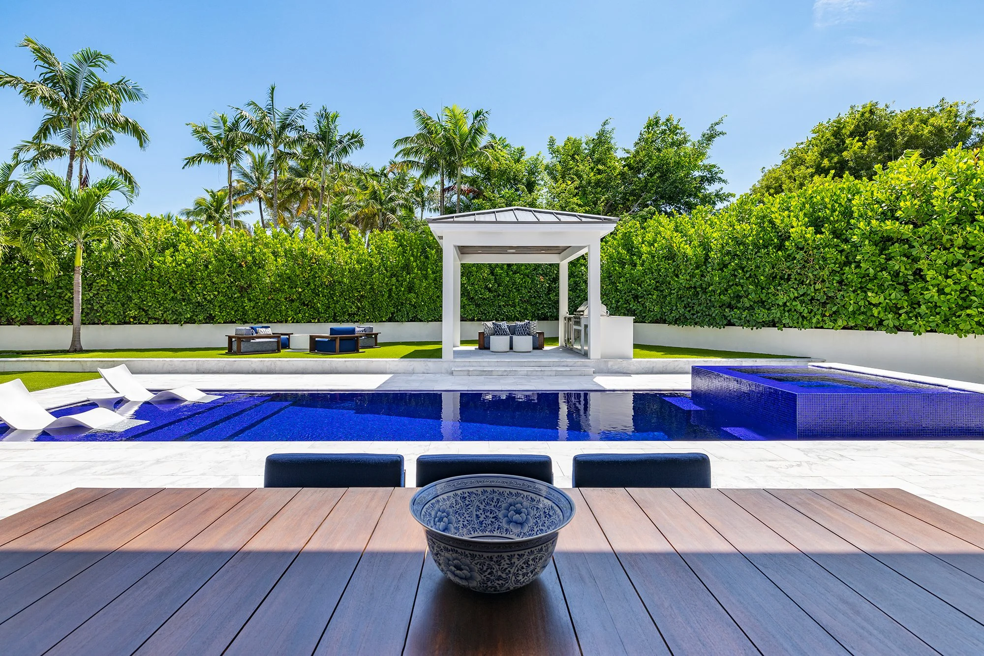 Backyard with a blue swimming pool, white lounge chairs, and a shaded seating area surrounded by lush green trees and palm trees under a clear blue sky.