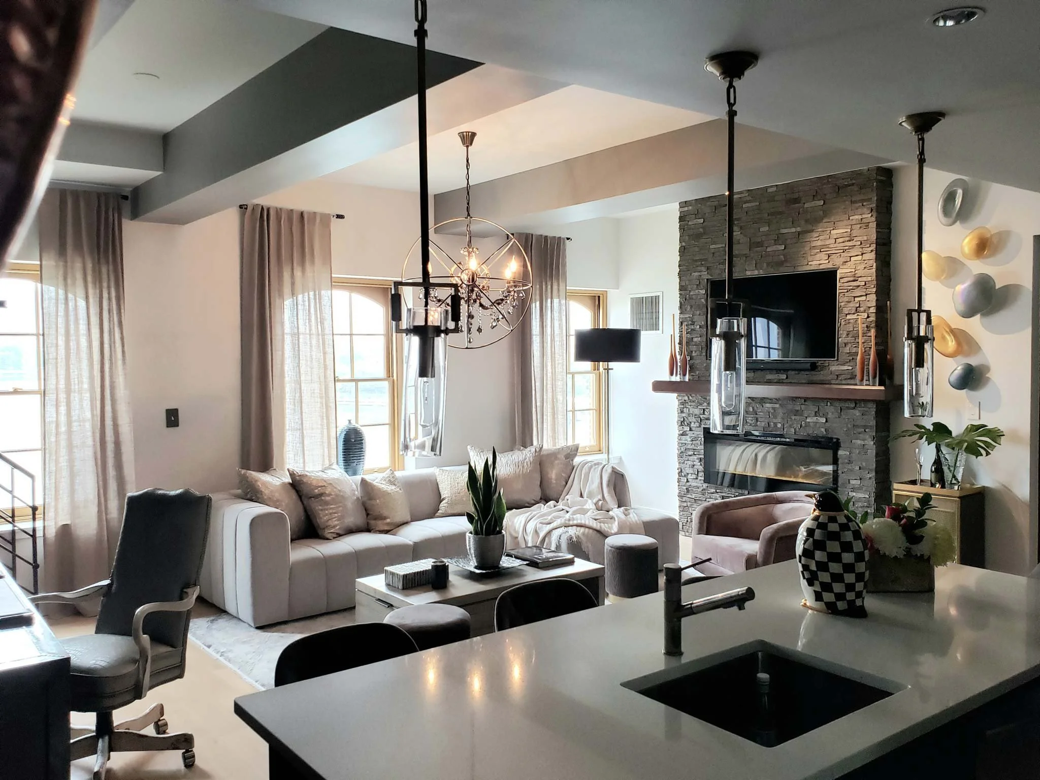 Living room with white sofa, beige curtains, stone fireplace, wall-mounted TV, modern lighting fixtures, and a kitchen island with a checkered vase.