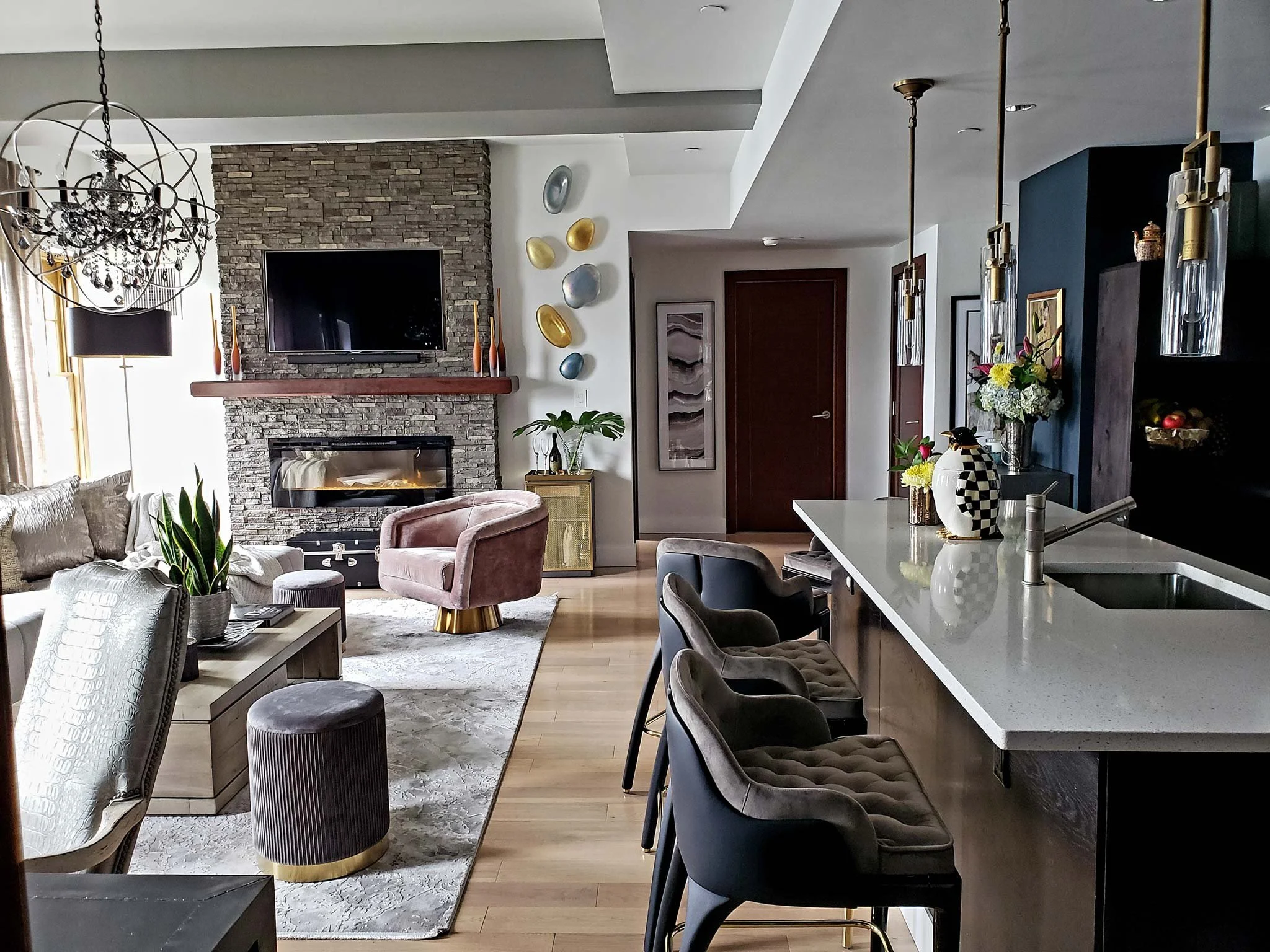 Modern living room and kitchen with stone fireplace, pink velvet chair, curved bar stools, pendant lights, artwork, floral arrangements, and hardwood floors.