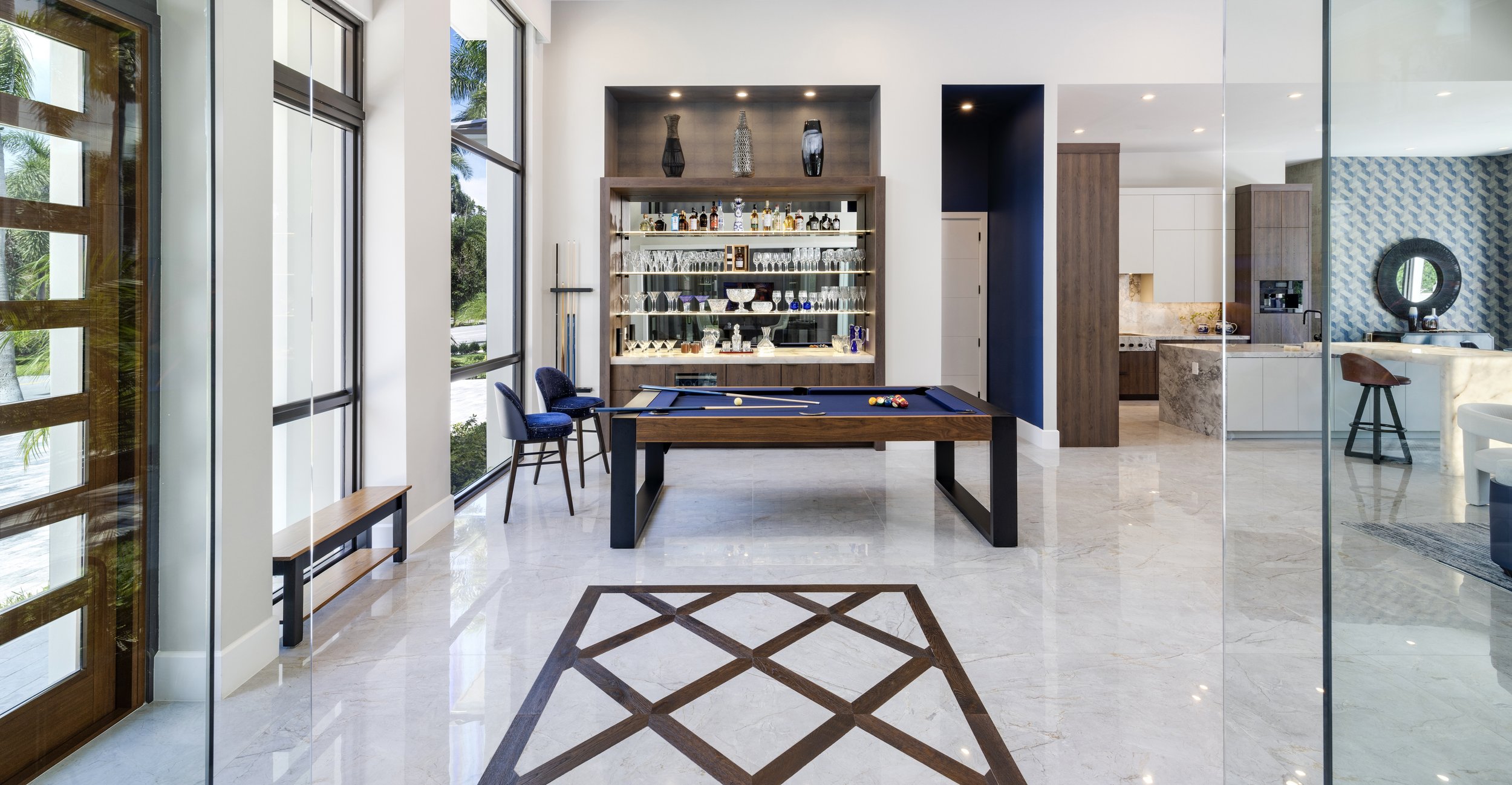 Interior of a modern house with a game room featuring a pool table, glass display cabinet with glassware and alcohol bottles, and seating area. The space has large windows with views of greenery outside, marble floors, and contemporary decor.