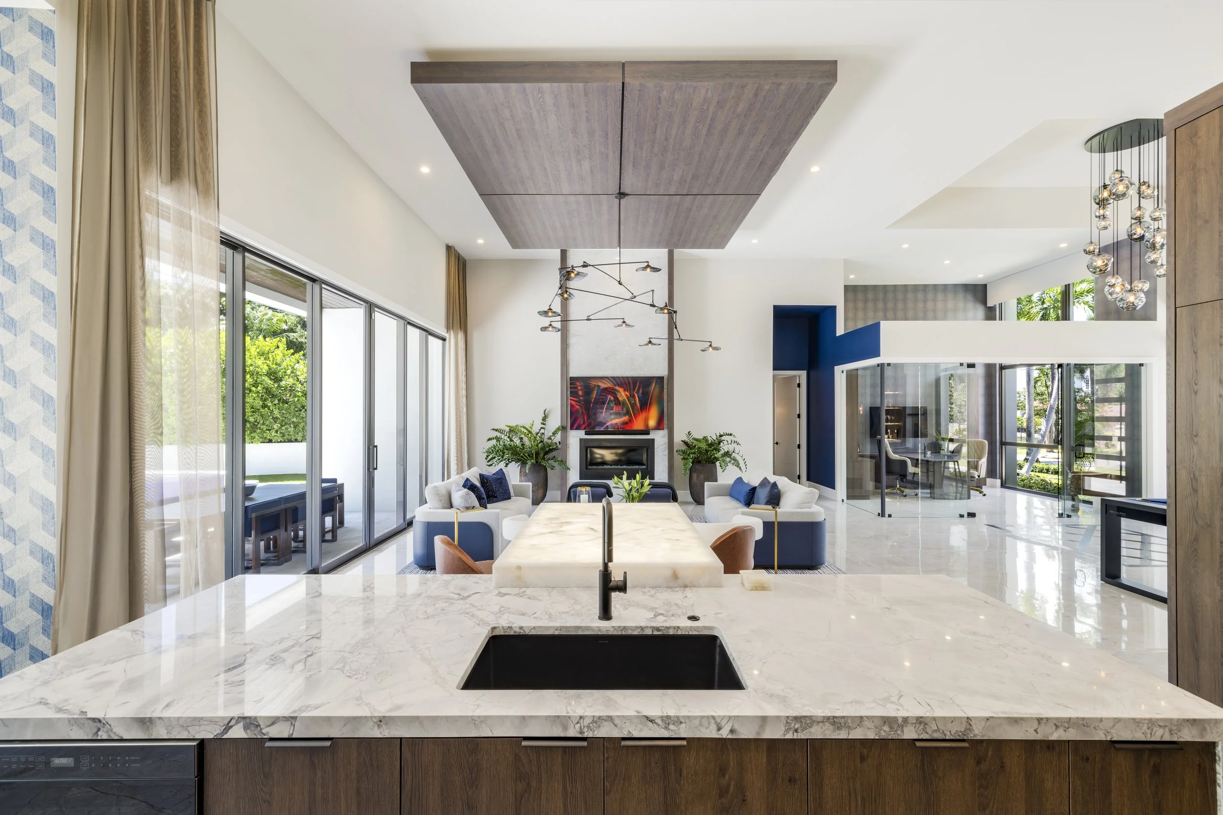 Open-concept living room and kitchen with marble island, modern furniture, large windows, and a fireplace.