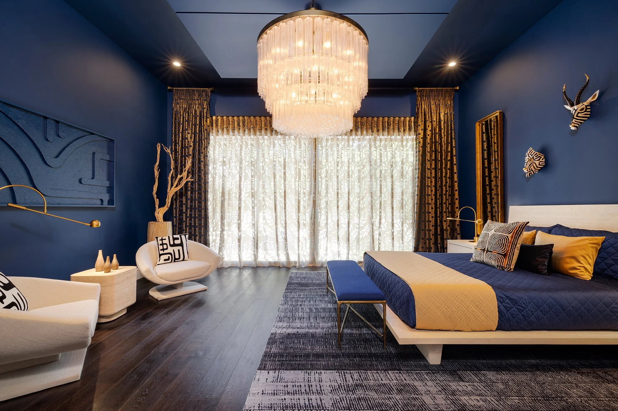 Modern bedroom with dark blue walls, white and yellow bedding on a low white bed, and black, white, and gold African animal wall art. Includes white lounge chairs, a black and gold bench, a large chandelier, patterned curtains, and dark hardwood floors.