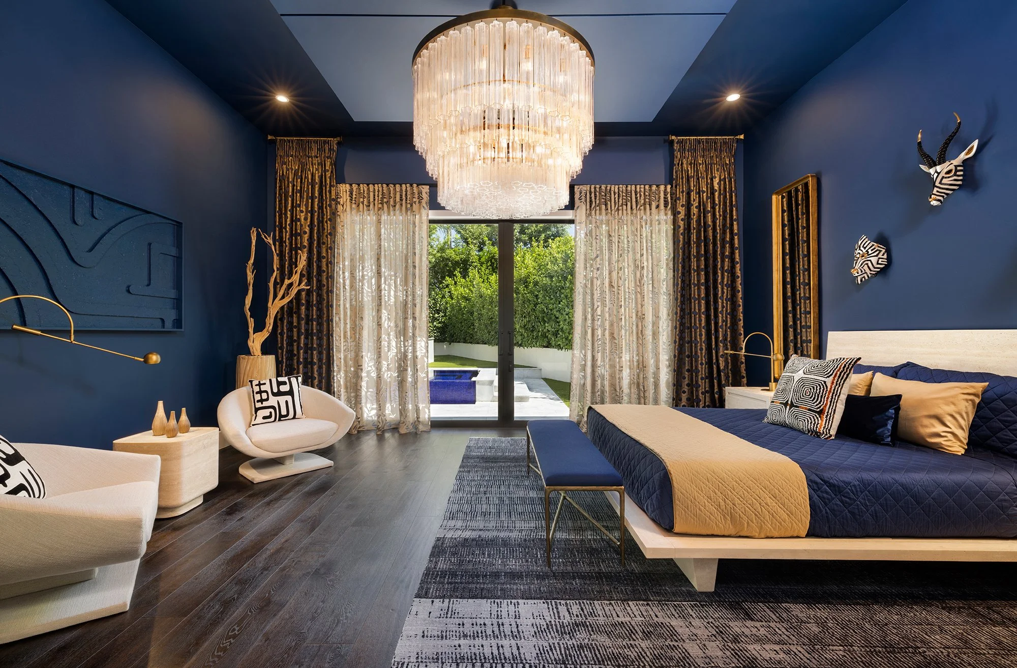 Modern bedroom with dark blue walls, a large chandelier, king-sized bed with a blue and beige quilt, decorative pillows, and a view of a backyard with pool and greenery through sliding glass doors.