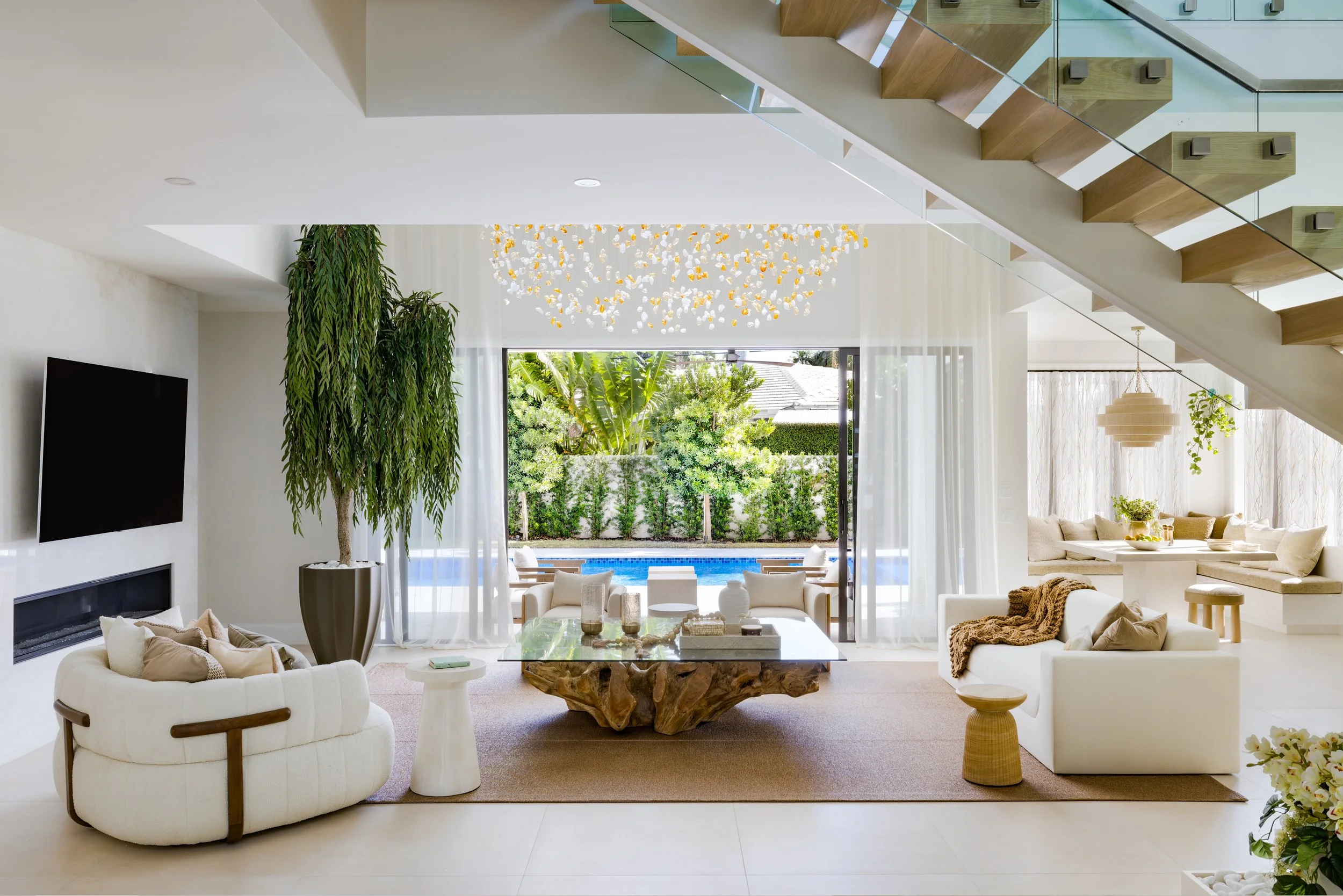 Modern living room with white furniture, a large potted plant, a stone coffee table, and view of a backyard with a pool and lush greenery.