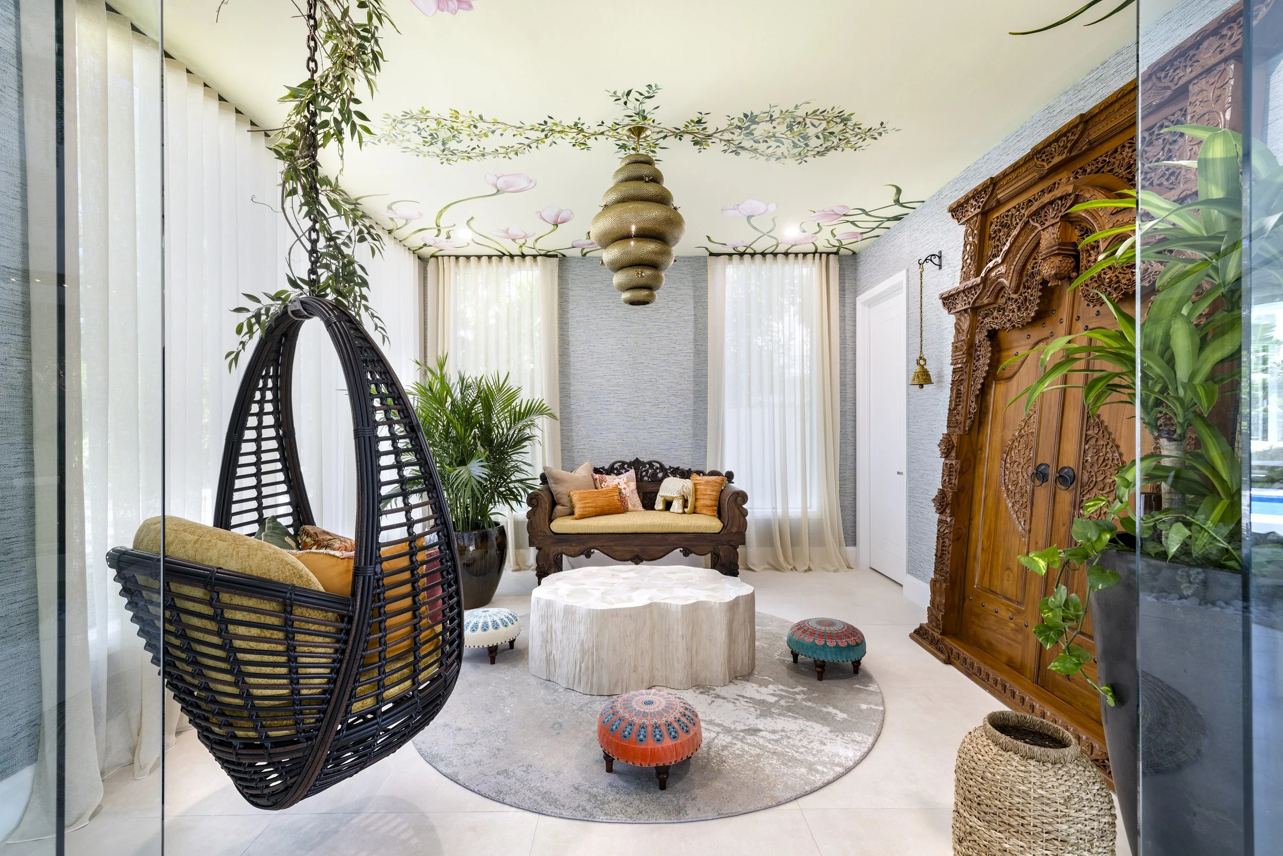 Bright living room with bohemian and eclectic decor, including a hanging egg chair, a carved wooden cabinet, potted plants, a round stone coffee table, and various colorful cushions and poufs, with light-colored curtains and ceiling painted with floral designs.