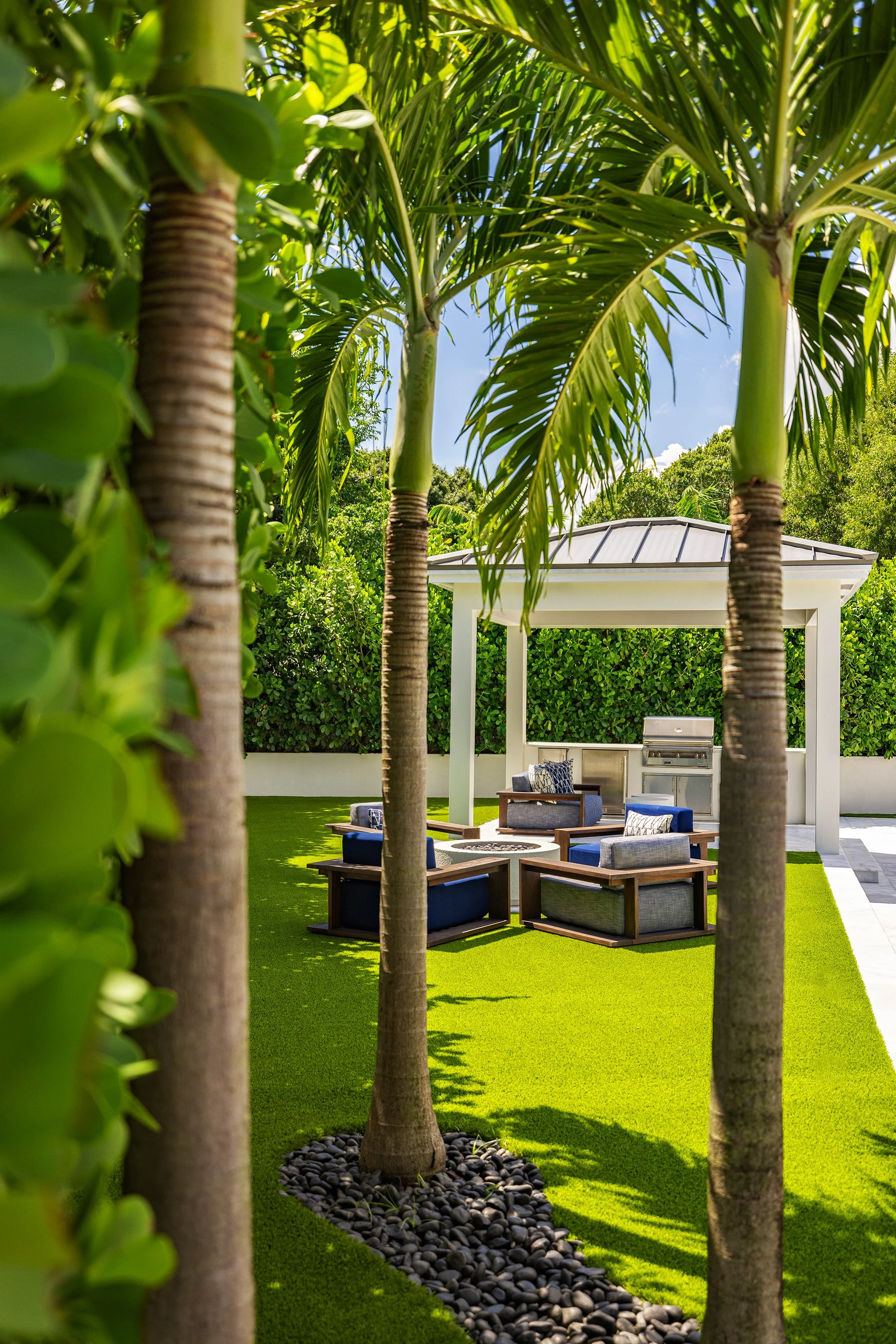 A backyard patio with a white pavilion, outdoor grill, and seating area with blue and gray chairs on a green lawn, surrounded by palm trees and bushes.