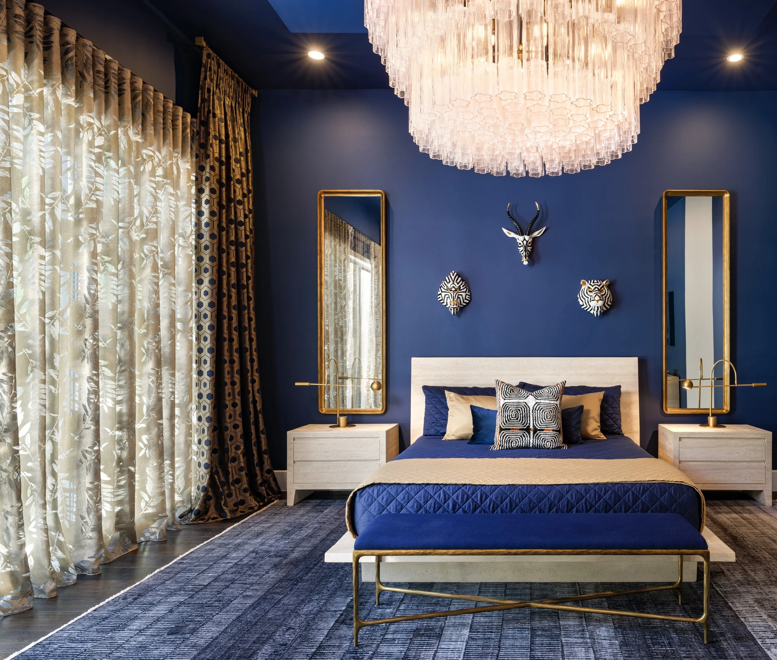 Modern bedroom with navy blue walls, gold accents, and African animal wall decor. Wooden furniture includes a bed with navy and beige bedding, matching nightstands with gold lamps, and two tall mirrors. Floor-to-ceiling curtains are patterned in earth tones. A large chandelier hangs from the ceiling.