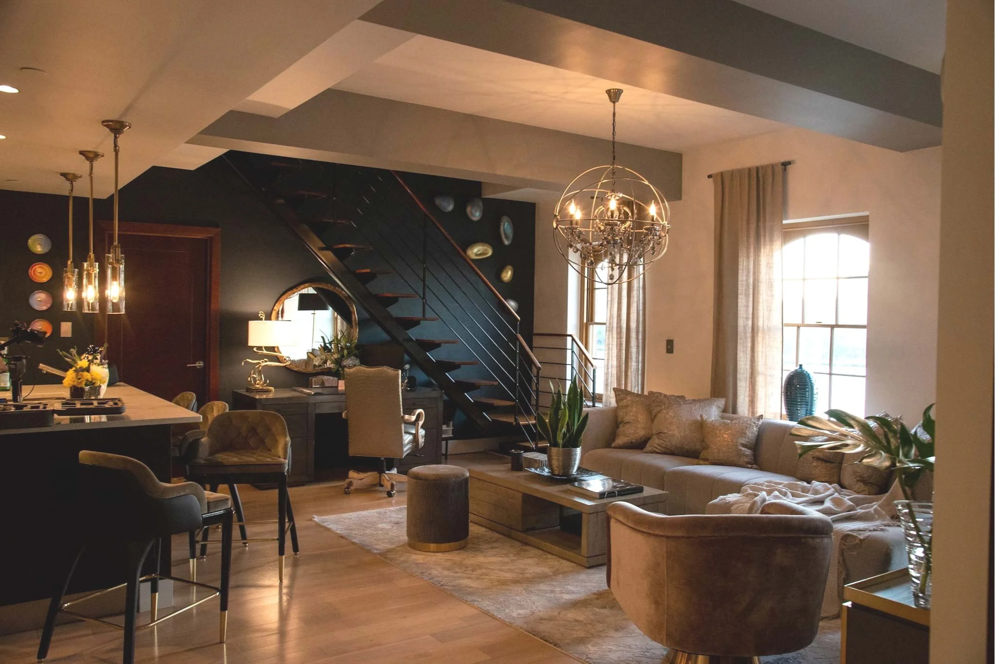 A cozy living room with a beige sectional sofa, a glass coffee table, and a chair. There are large windows with curtains, a chandelier hanging from the ceiling, and a wooden staircase leading upstairs. Decor includes potted plants, a mirror, and wall art.