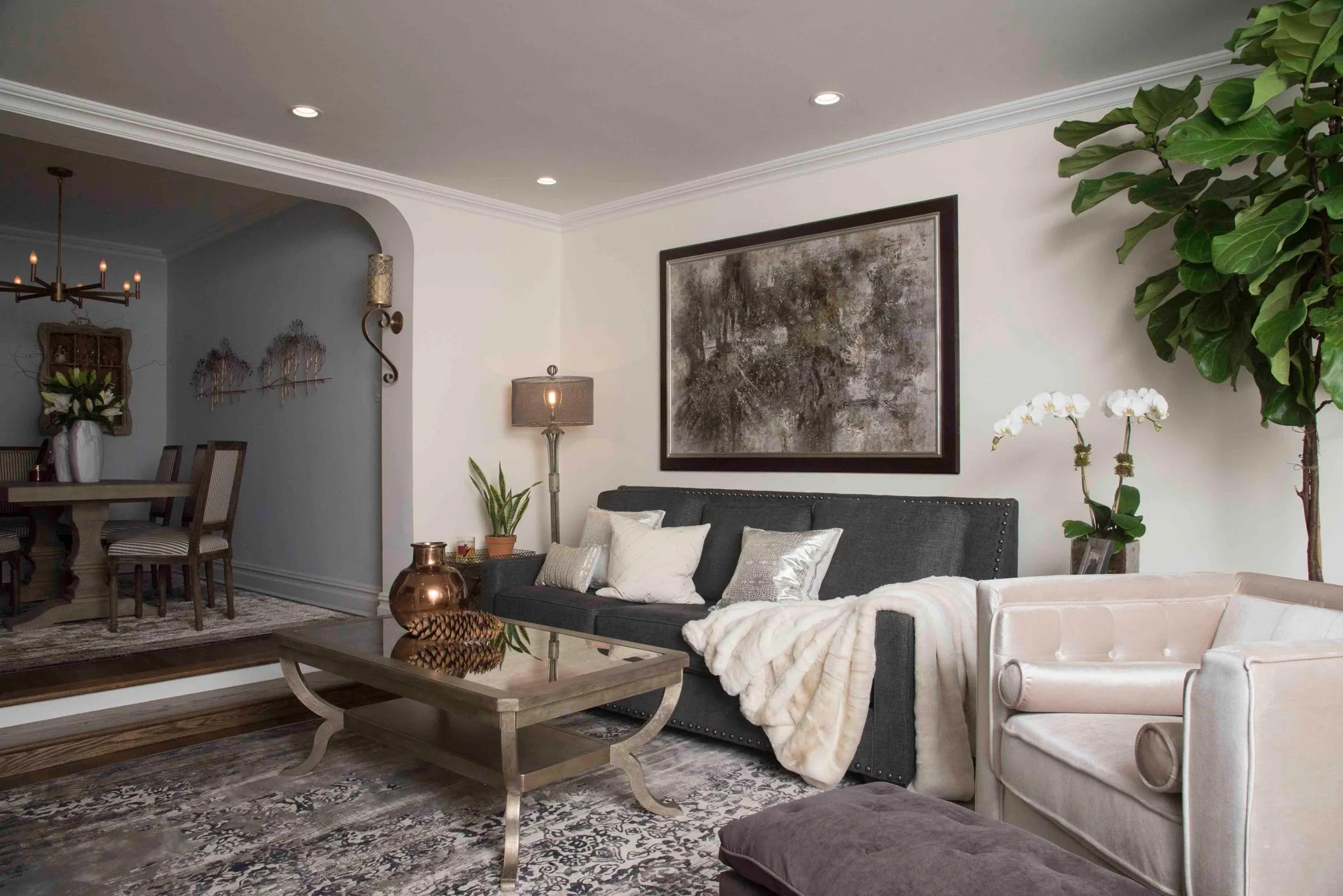 Living room with a dark gray sofa, white armchair, and a glass-topped coffee table. Decor includes plants, a large abstract painting, and decorative pillows, with a dining area visible through an arch.