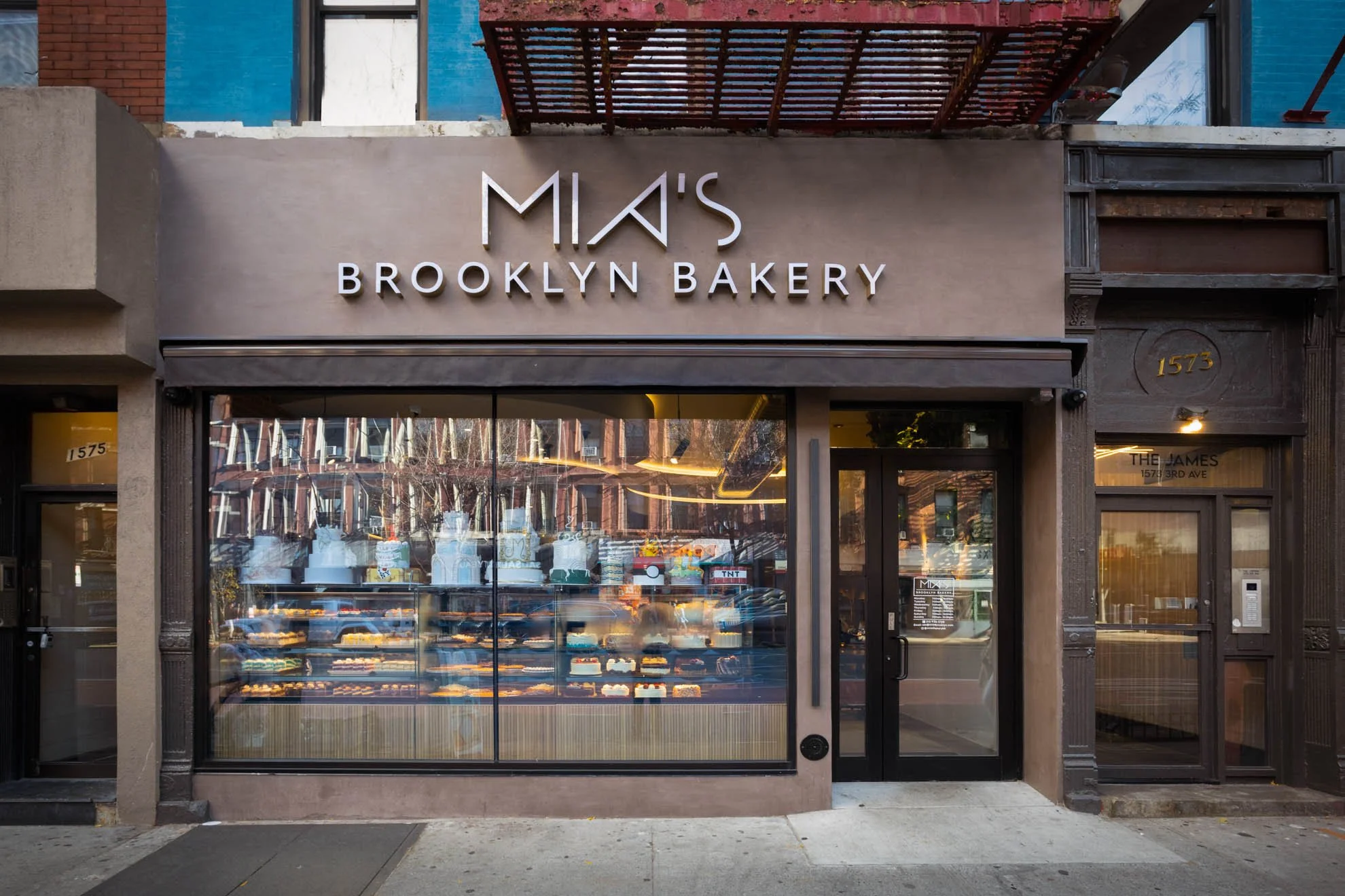 storefront of Mia's Brooklyn Bakery with a large glass window displaying baked goods inside and a beige exterior wall with the business name in large metal letters.