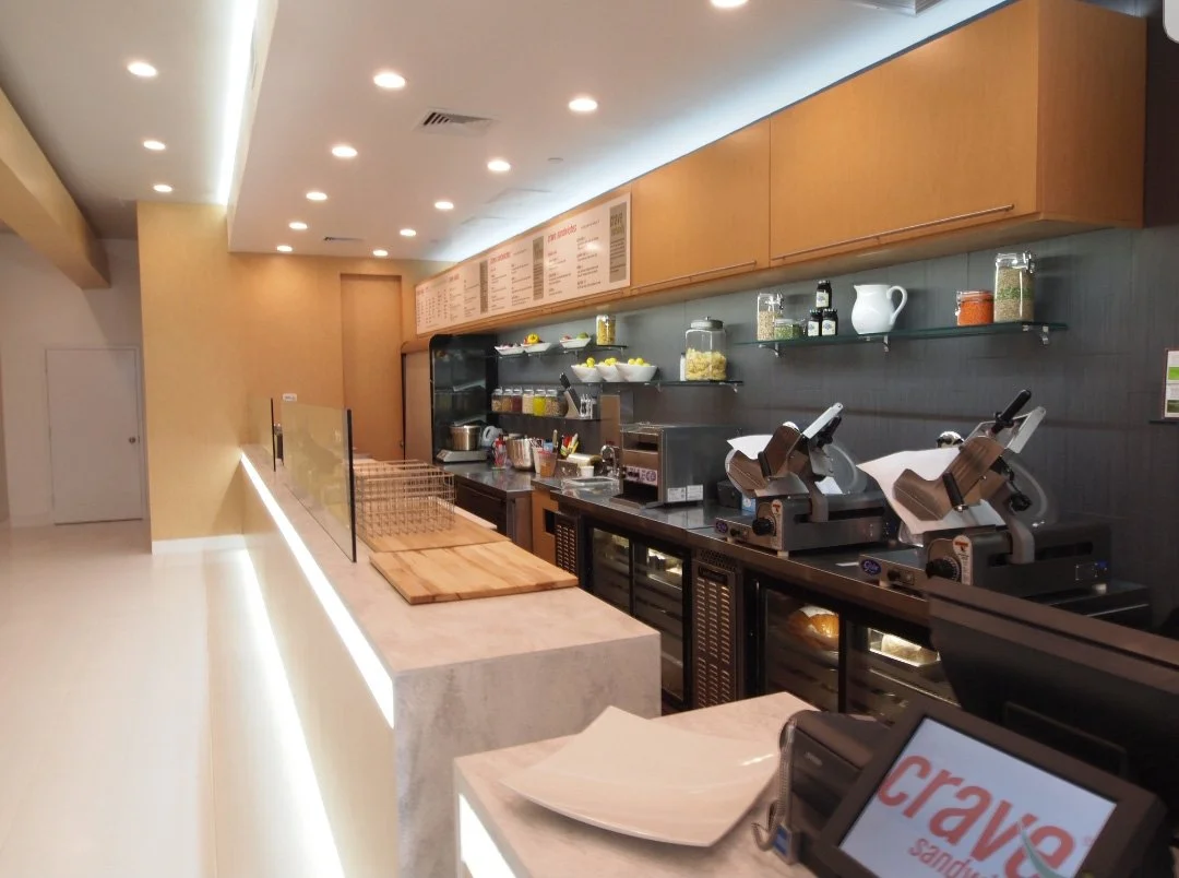 A modern cafe counter with pizza slicers, chopping boards, and refrigerated display cases, behind brown wood cabinets, with a menu on the wall.