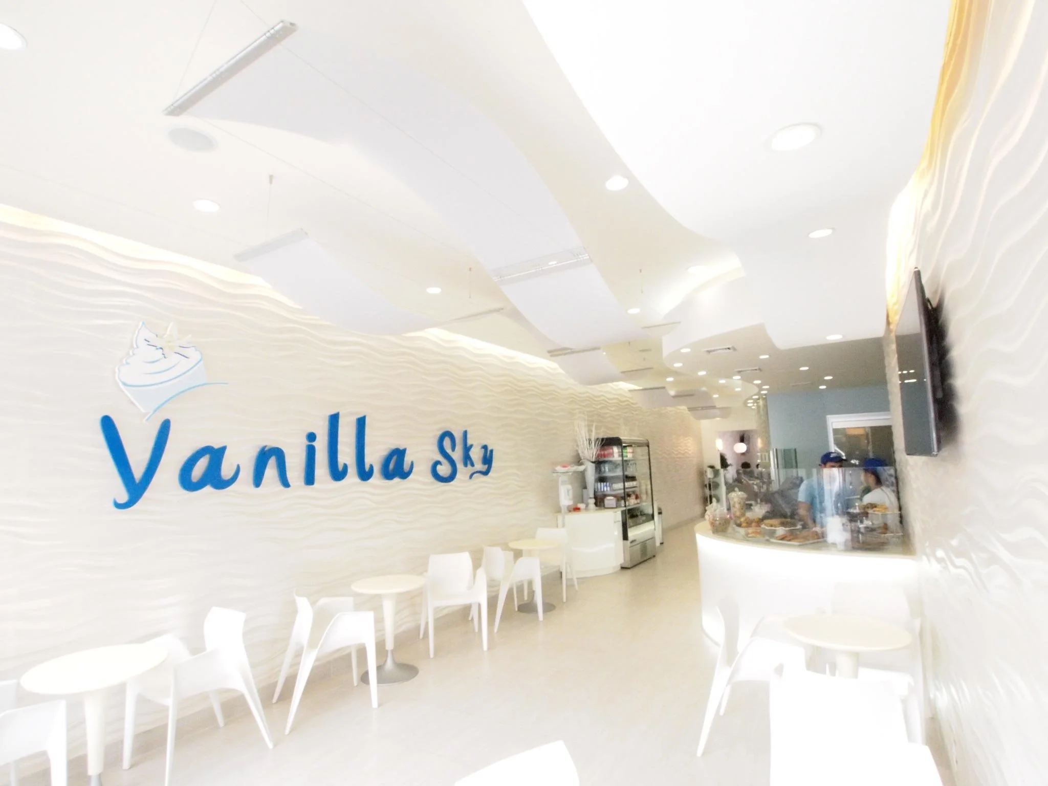 Interior of Vanilla Sky ice cream shop with white walls, minimalist white tables and chairs, a counter with staff, and a large screen on the wall.
