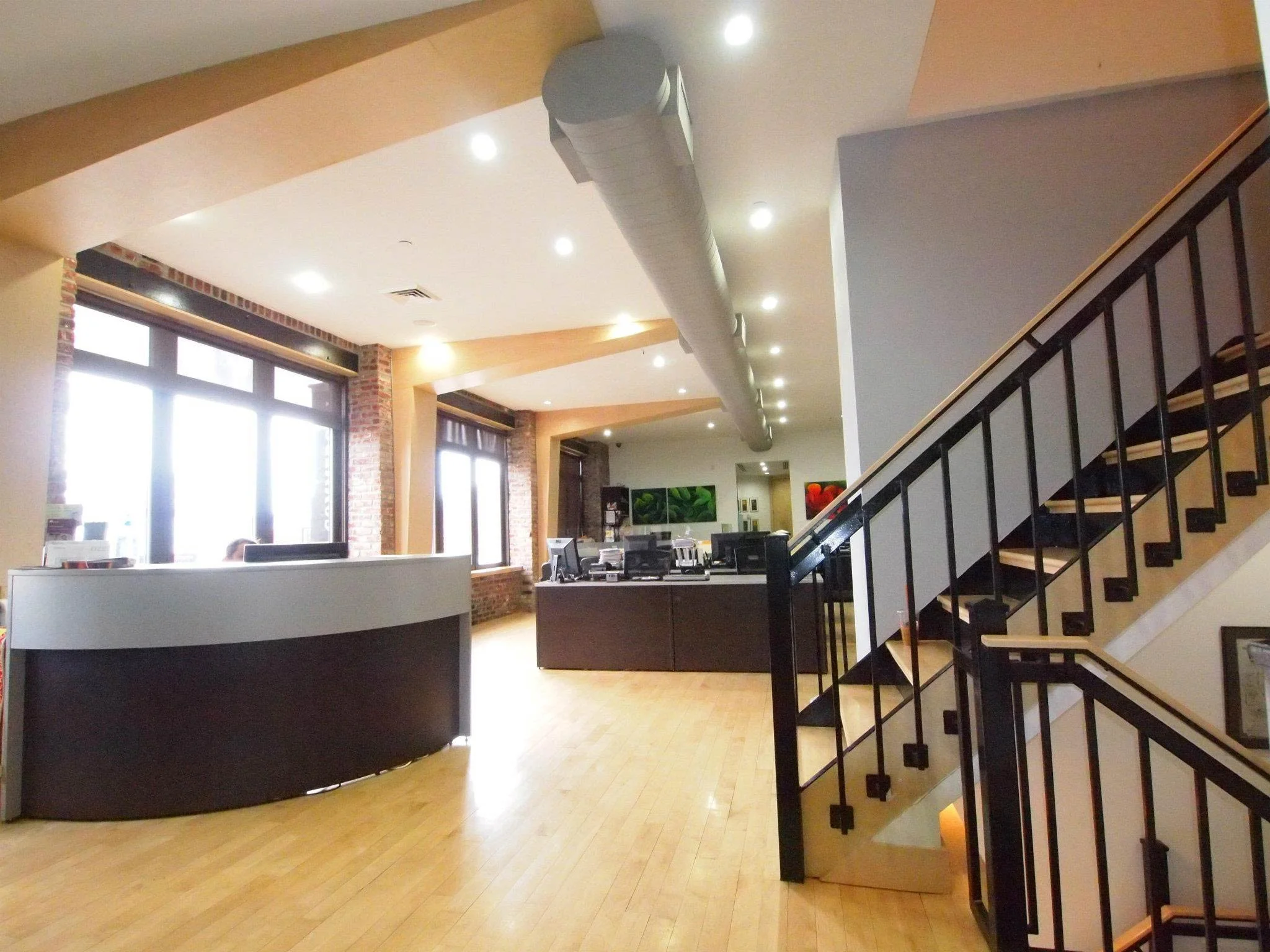 Interior of a modern office or reception area with large windows, brick accents, a curved reception desk, a staircase with black railing, and a row of computer workstations.