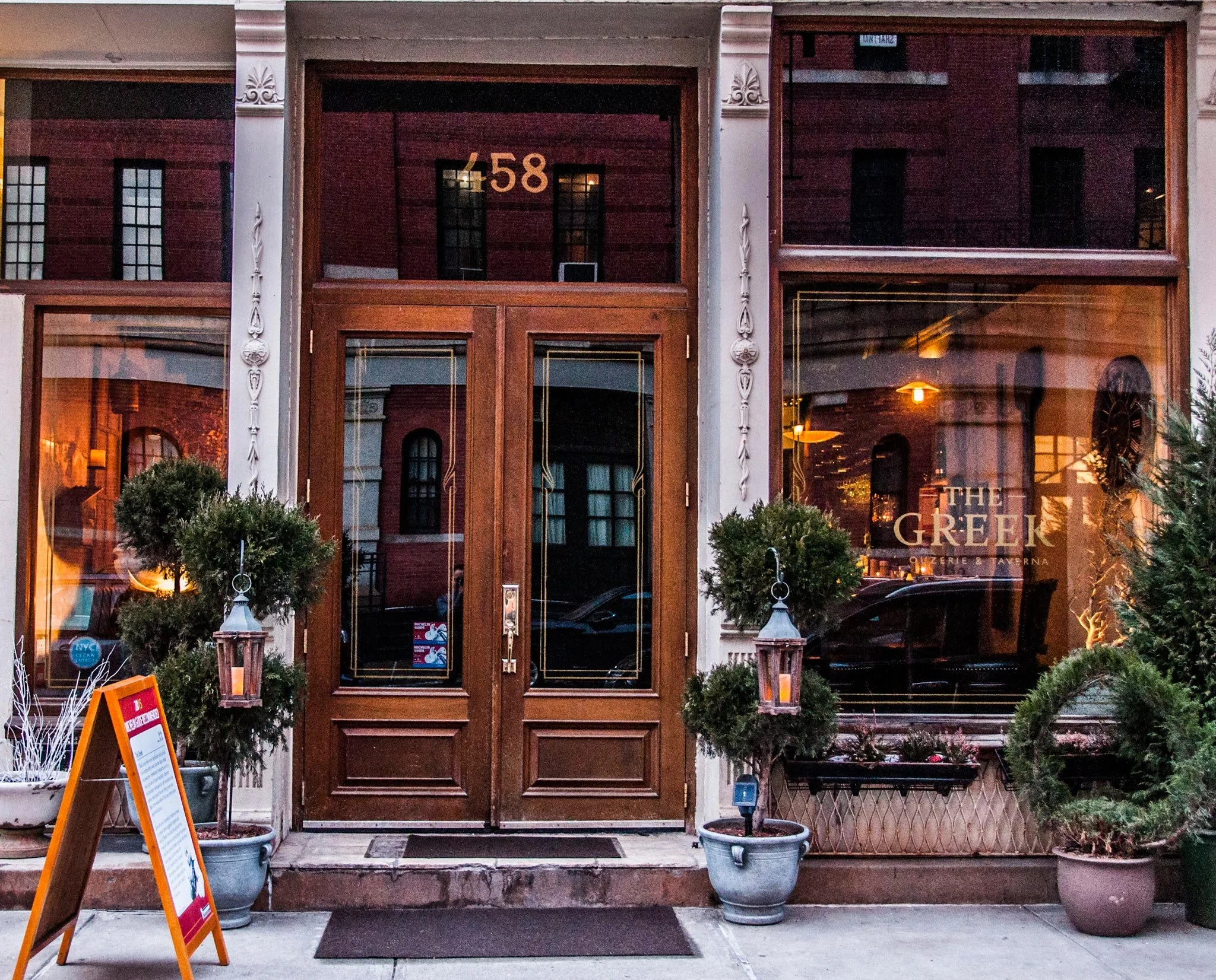 The entrance to a restaurant called The Greek, with wooden doors, potted bushes, and lanterns, and a signboard outside.