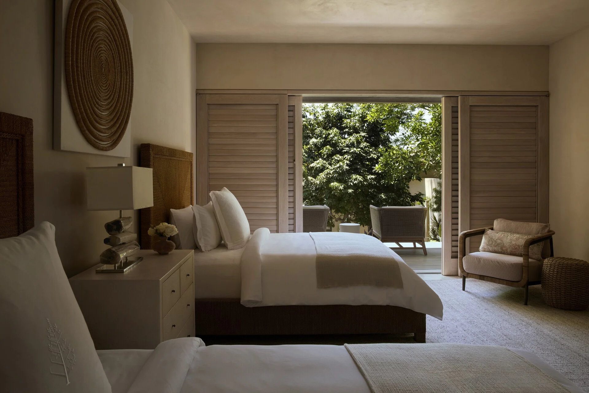 Hotel room with two beds, sliding doors open to a balcony with outdoor chairs and lush green trees outside.