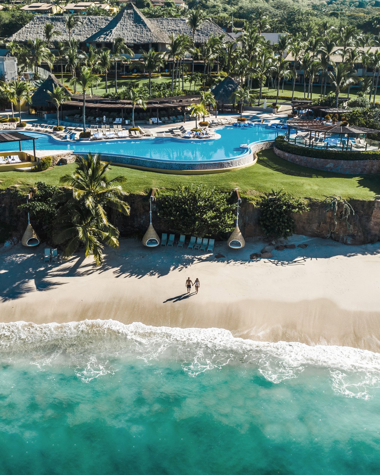 Aerial view of a tropical resort with a large swimming pool, surrounded by palm trees, lounge chairs, and thatched-roof buildings. In the foreground, a couple is walking along the sandy beach near the ocean.