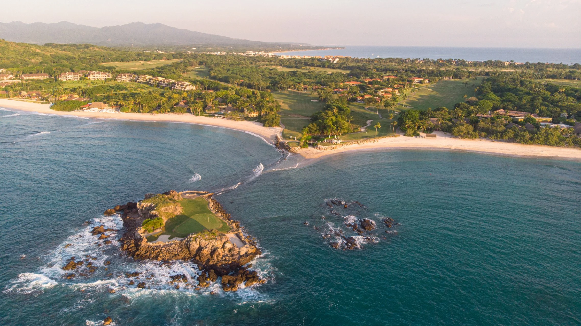 Aerial view of a coastal area with beaches, ocean, rocky outcroppings, a small golf course on rocks, residential houses, green hills, and distant mountains.