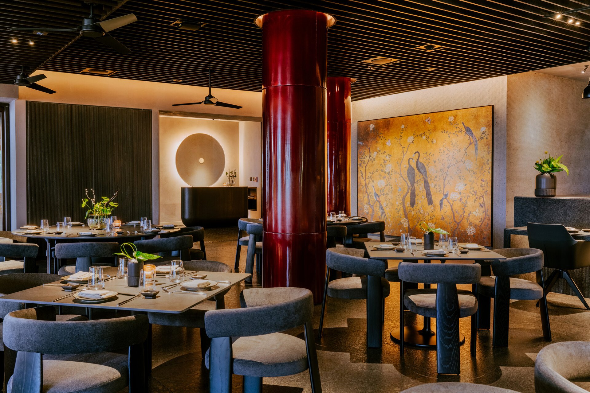 Elegant restaurant interior with multiple round tables set with glasses, plates, and cutlery, surrounded by cushioned chairs, featuring red glossy columns, a large decorative Asian-inspired mural of birds, and modern minimalist decor with subtle ligh