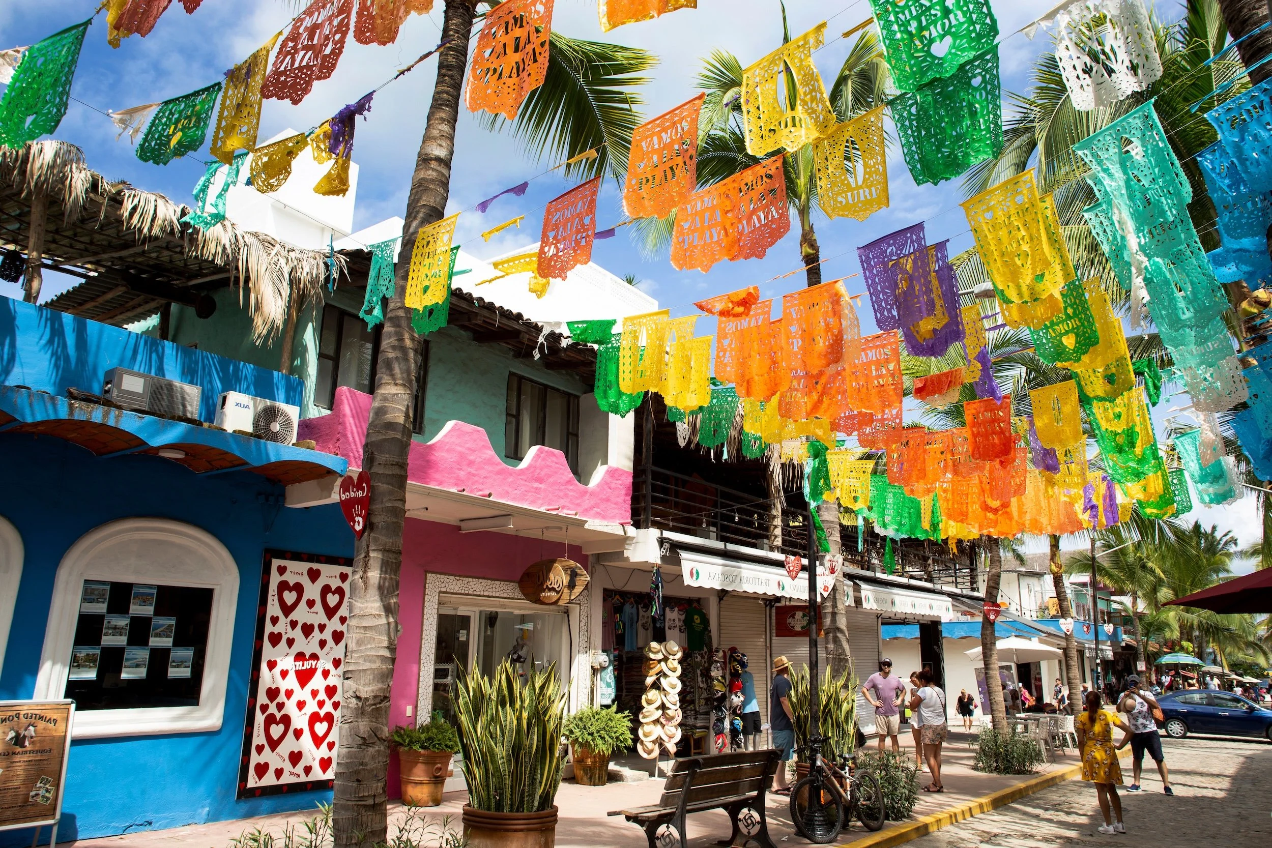 Street scene with colorful papel picado decorations hanging above, palm trees, and shops with vibrant facades on a sunny day. People are walking and relaxing on the sidewalk.