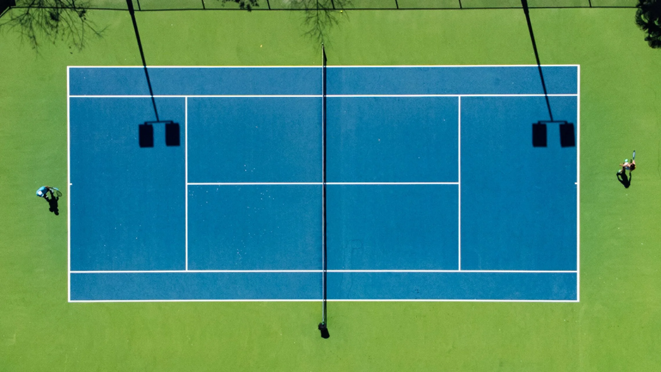 Aerial view of a blue tennis court with surrounds of green grass, two people at opposite ends, and overhead lighting.