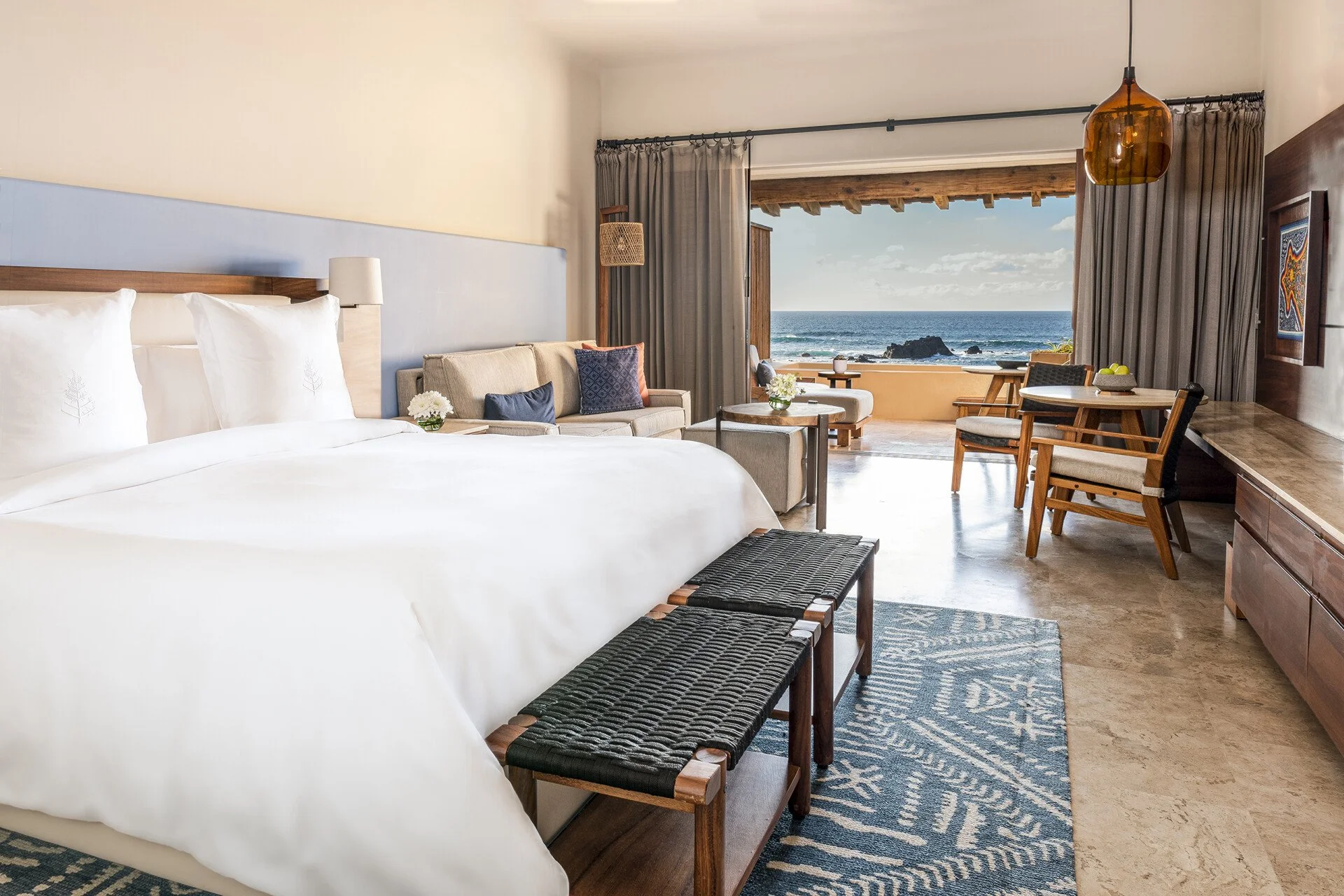 A hotel room with a large bed, sofa, and a view of the ocean through an open balcony with curtains. There are chairs, a table, and decorative items in the room.