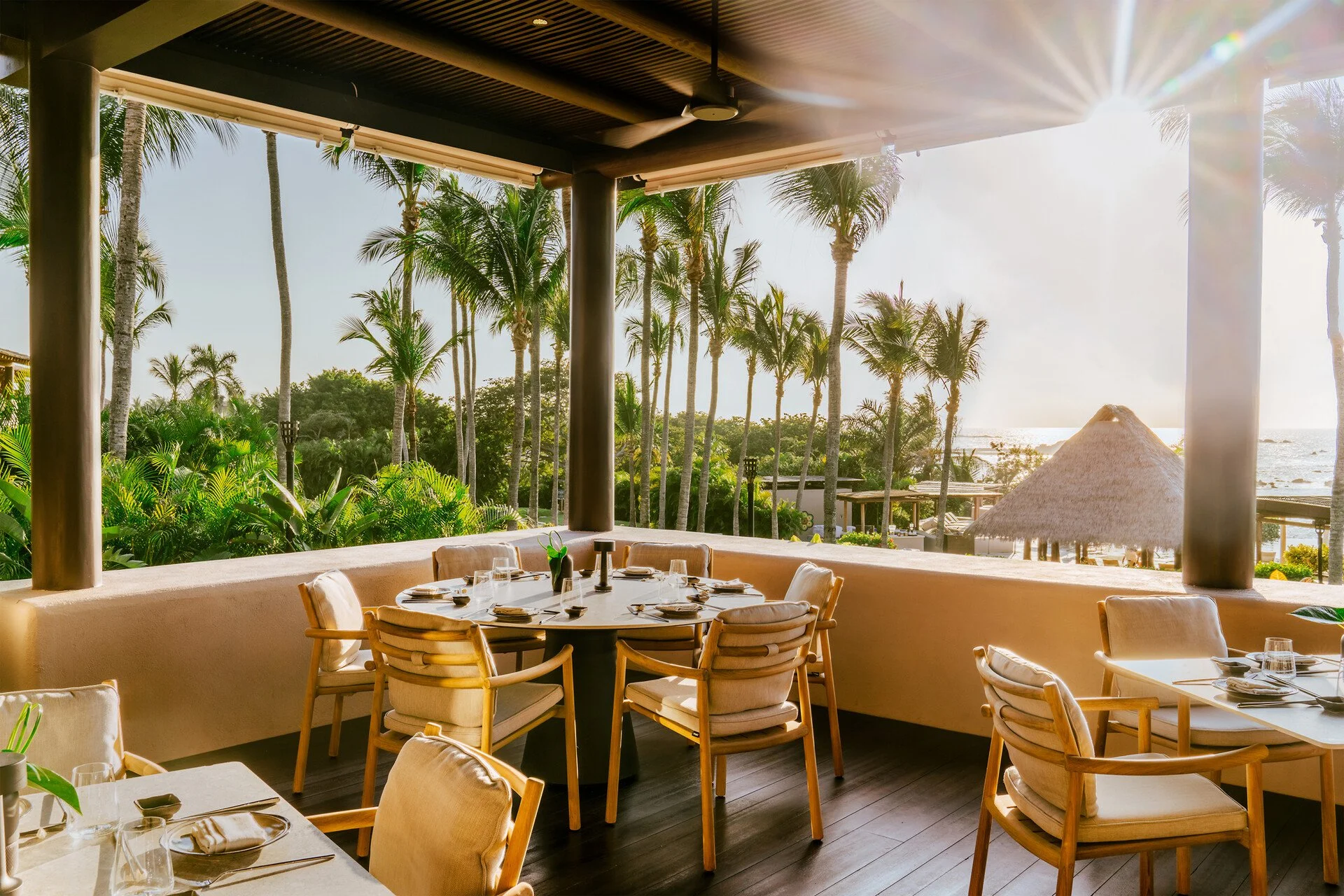 A tropical outdoor dining area with a view of palm trees, greenery, a thatched roof hut, and the ocean in the background, illuminated by sunlight.