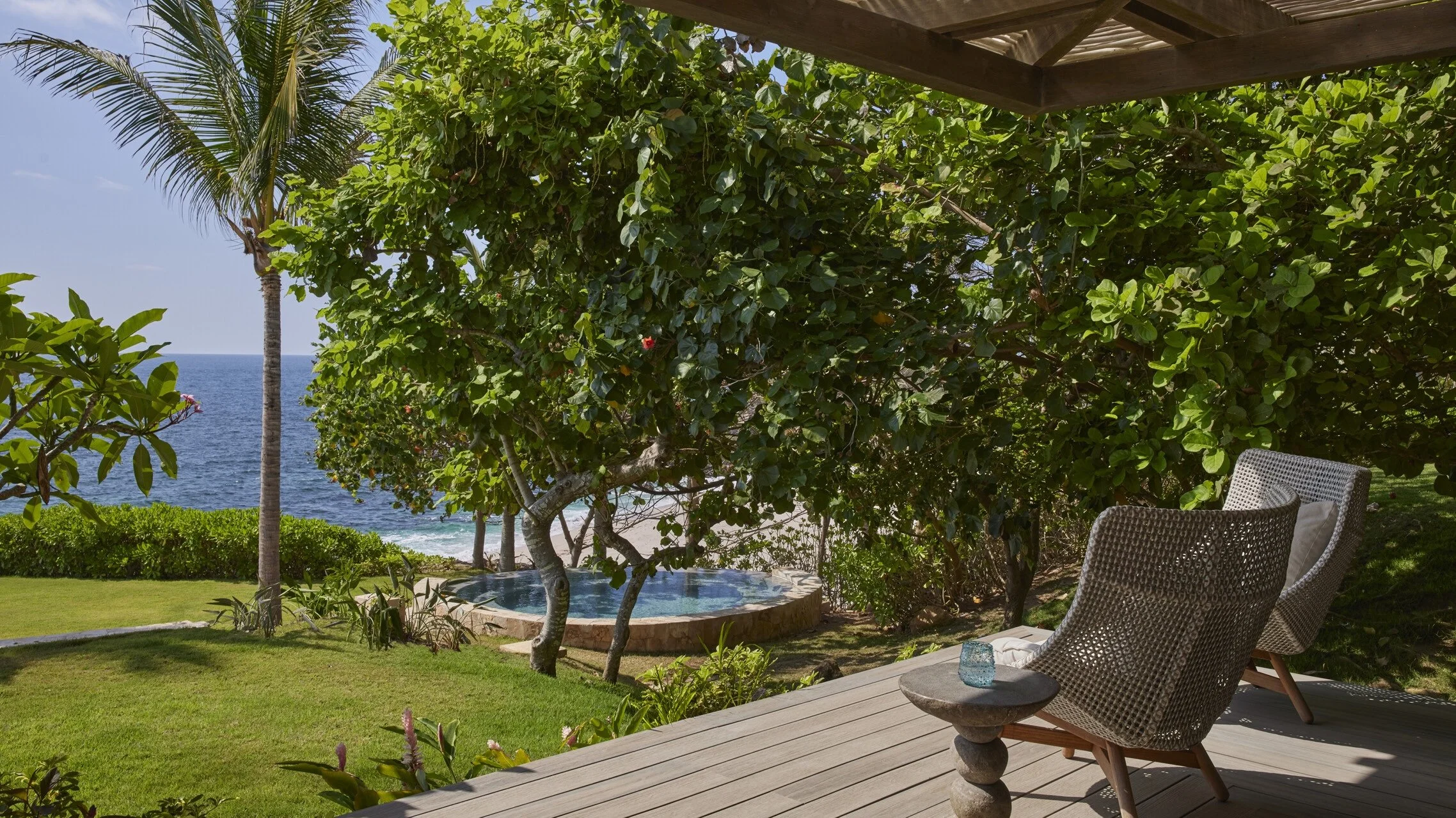 A tropical outdoor scene with a wooden deck and two wicker chairs facing the ocean, lush green trees, a small pool, a palm tree, and the sea in the background.