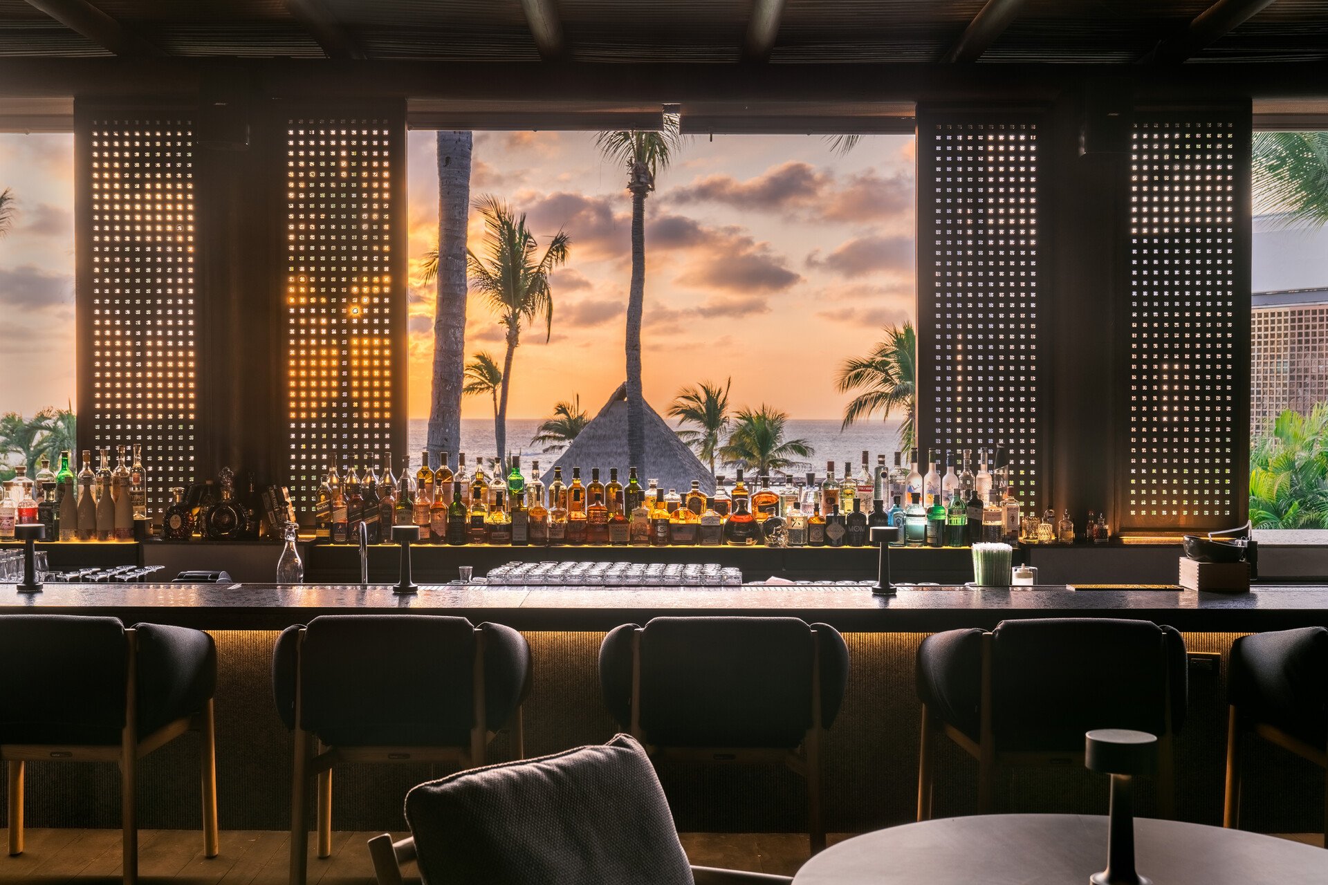 Bar with bottles of alcohol and a view of palm trees and a sunset over the ocean in the background.