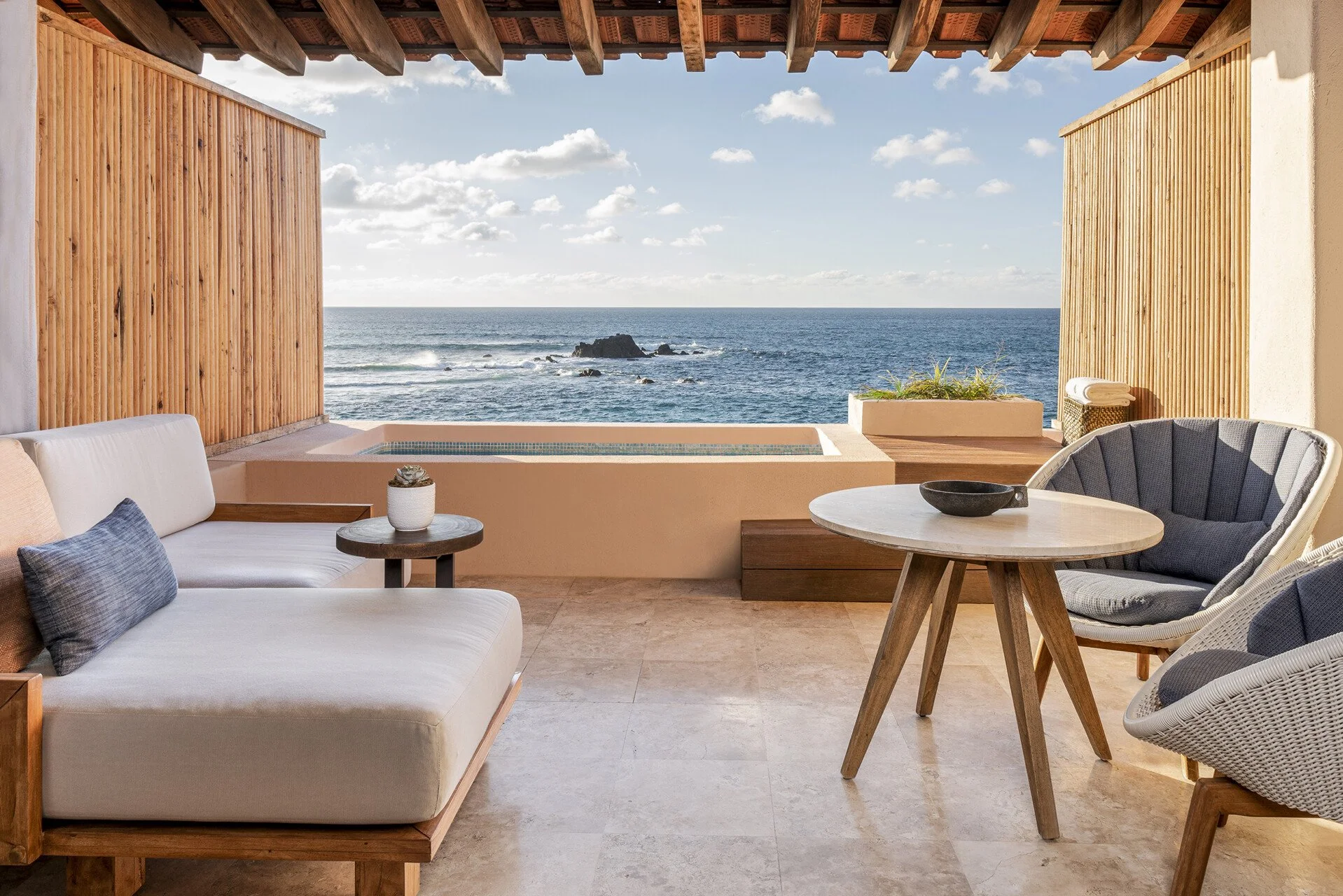 Beachside patio with white sofa, wooden side table, round dining table, two cushioned chairs, and ocean view with rocks and clouds.