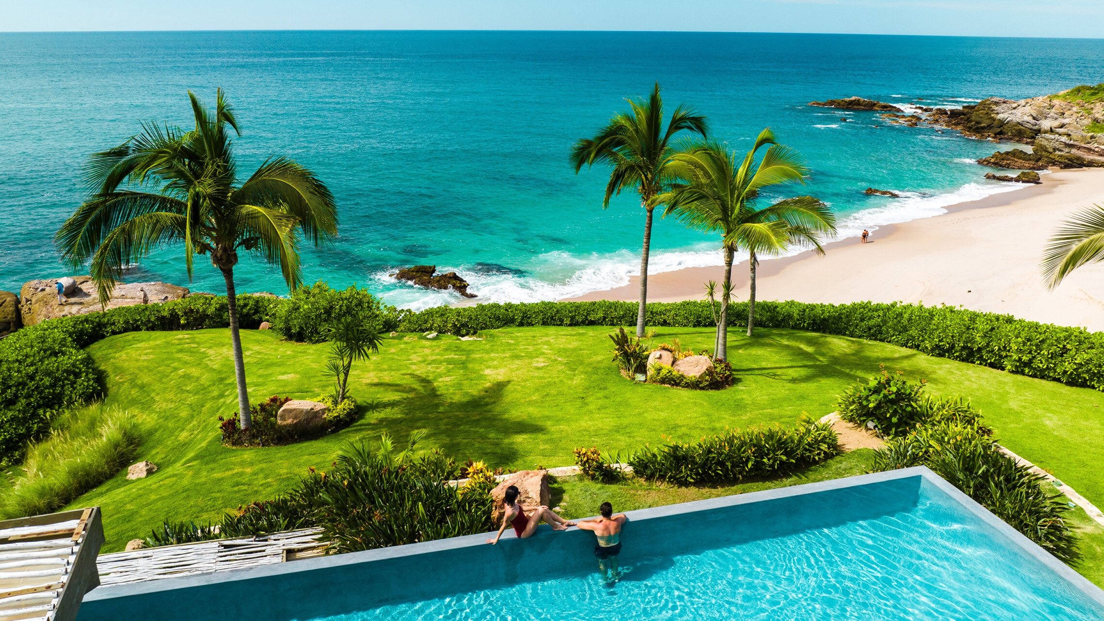 Two women sitting on the edge of a swimming pool overlooking a tropical beach with palm trees, green lawn, and ocean waves.