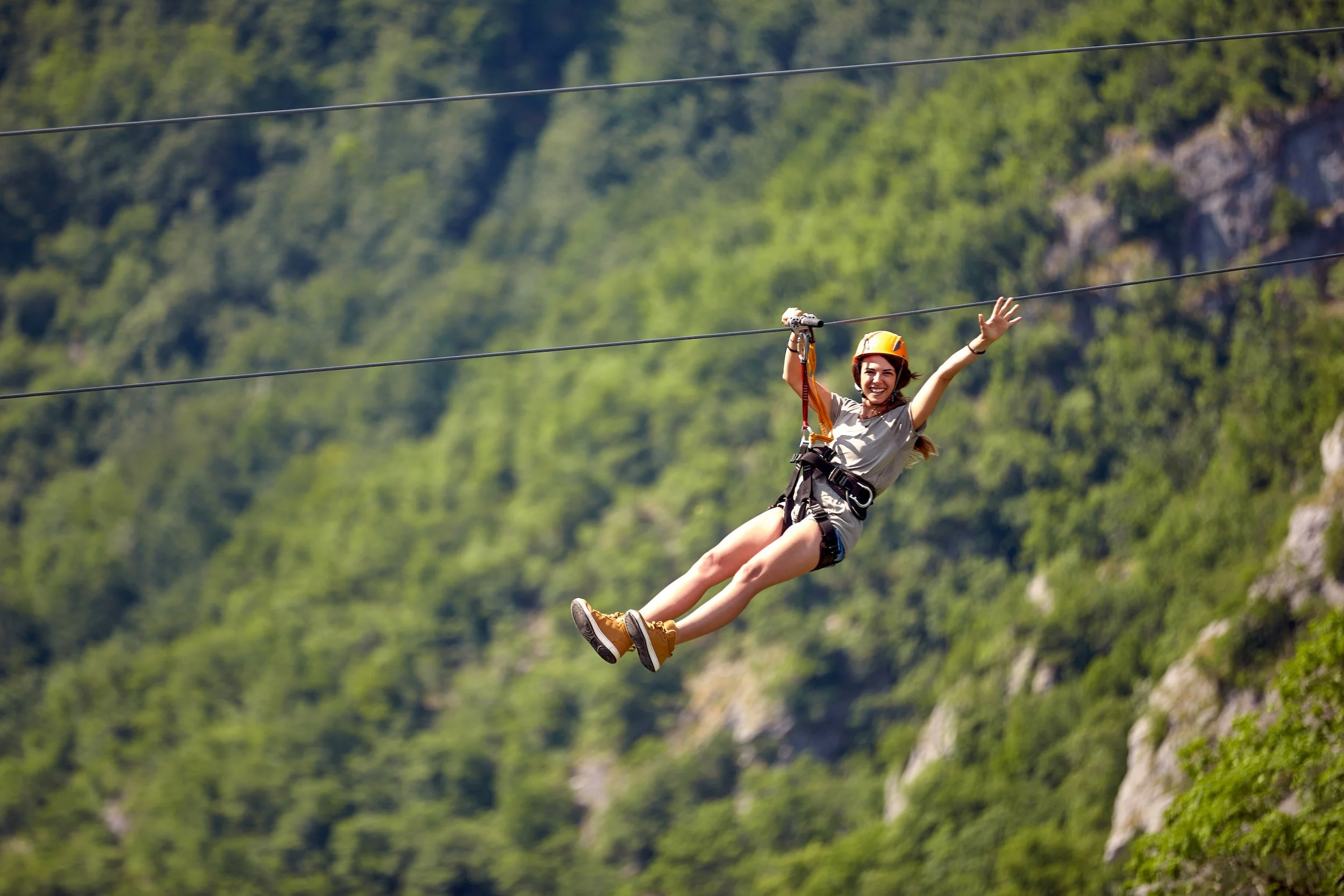 A woman wearing a helmet and harness riding a zip line over a green, mountainous landscape, smiling and waving.